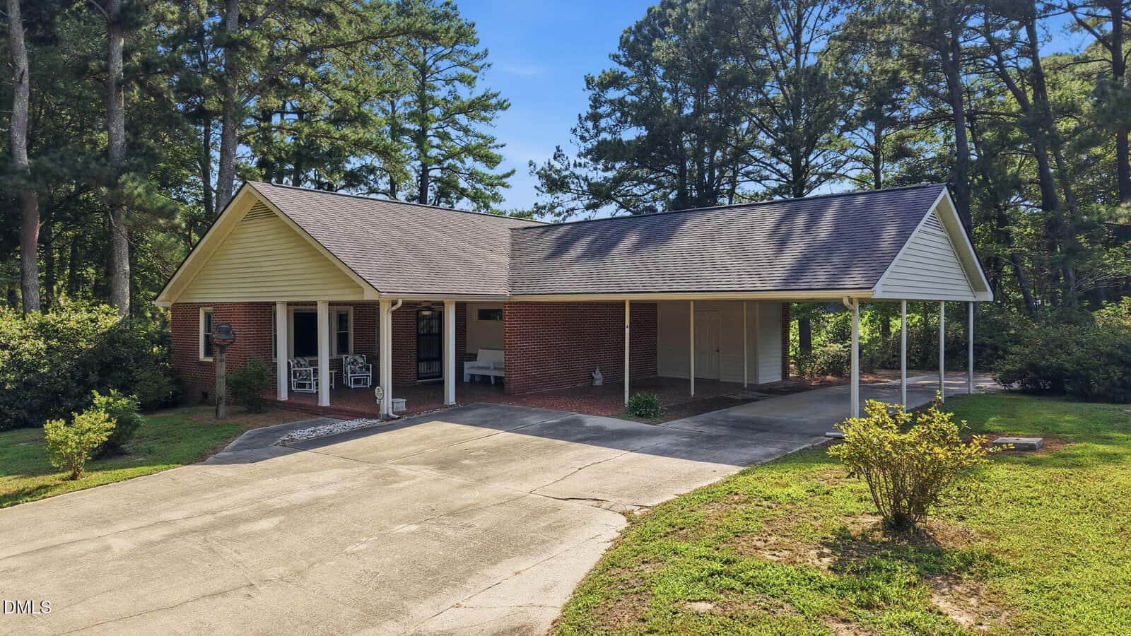 A one-story brick house with a gable roof, covered porch, and attached carport is nestled at 69 Dyking Road in Louisburg. The driveway curves toward the carport, while grass, shrubs, and tall pine trees surround the property.