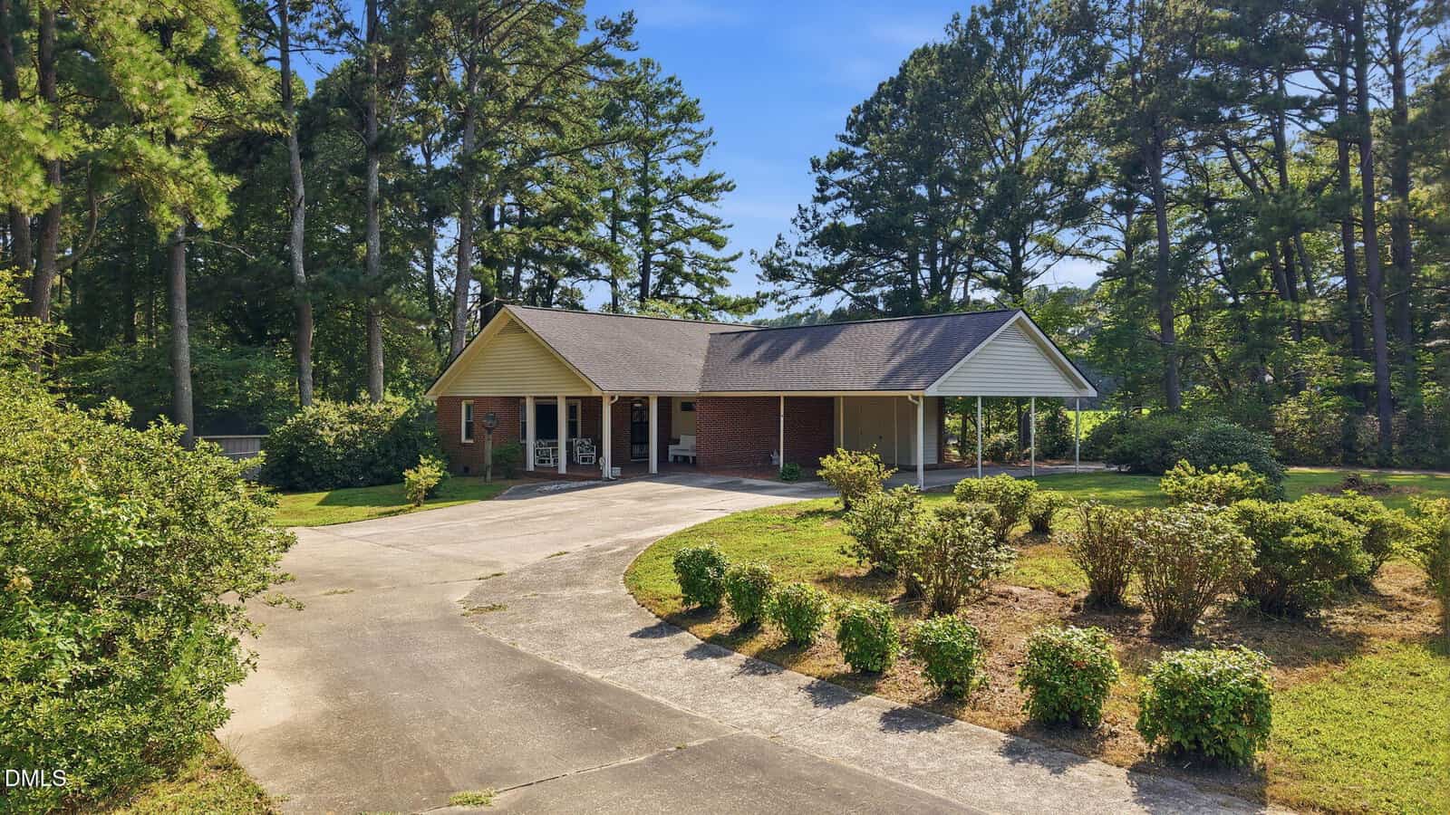 One-story brick house with a covered front porch and carport, surrounded by bushes and trees. A curved driveway leads to the house, and the lawn is well-maintained under a clear blue sky.