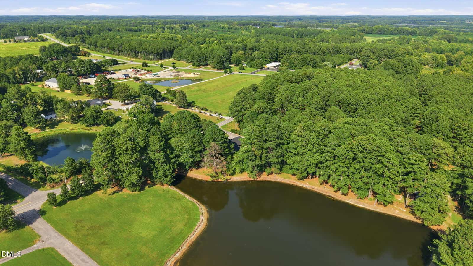 Aerial view of a rural area with a pond in the foreground, a mix of wooded areas and open fields, scattered houses, and a road running through the landscape under a blue sky.