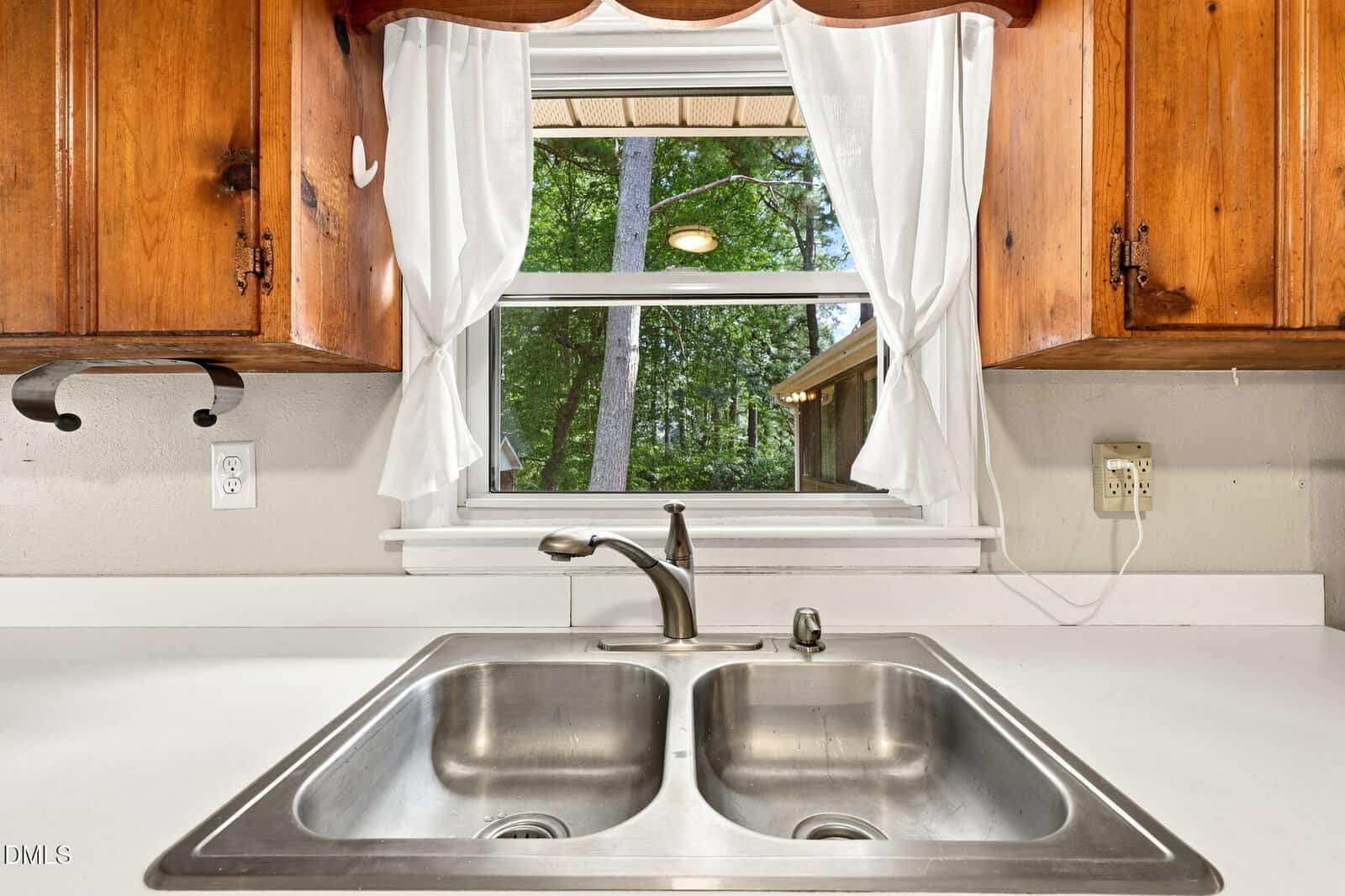 A stainless steel double kitchen sink with a faucet sits below a window with white curtains, offering a view of trees outside. Wooden cabinets frame the window.