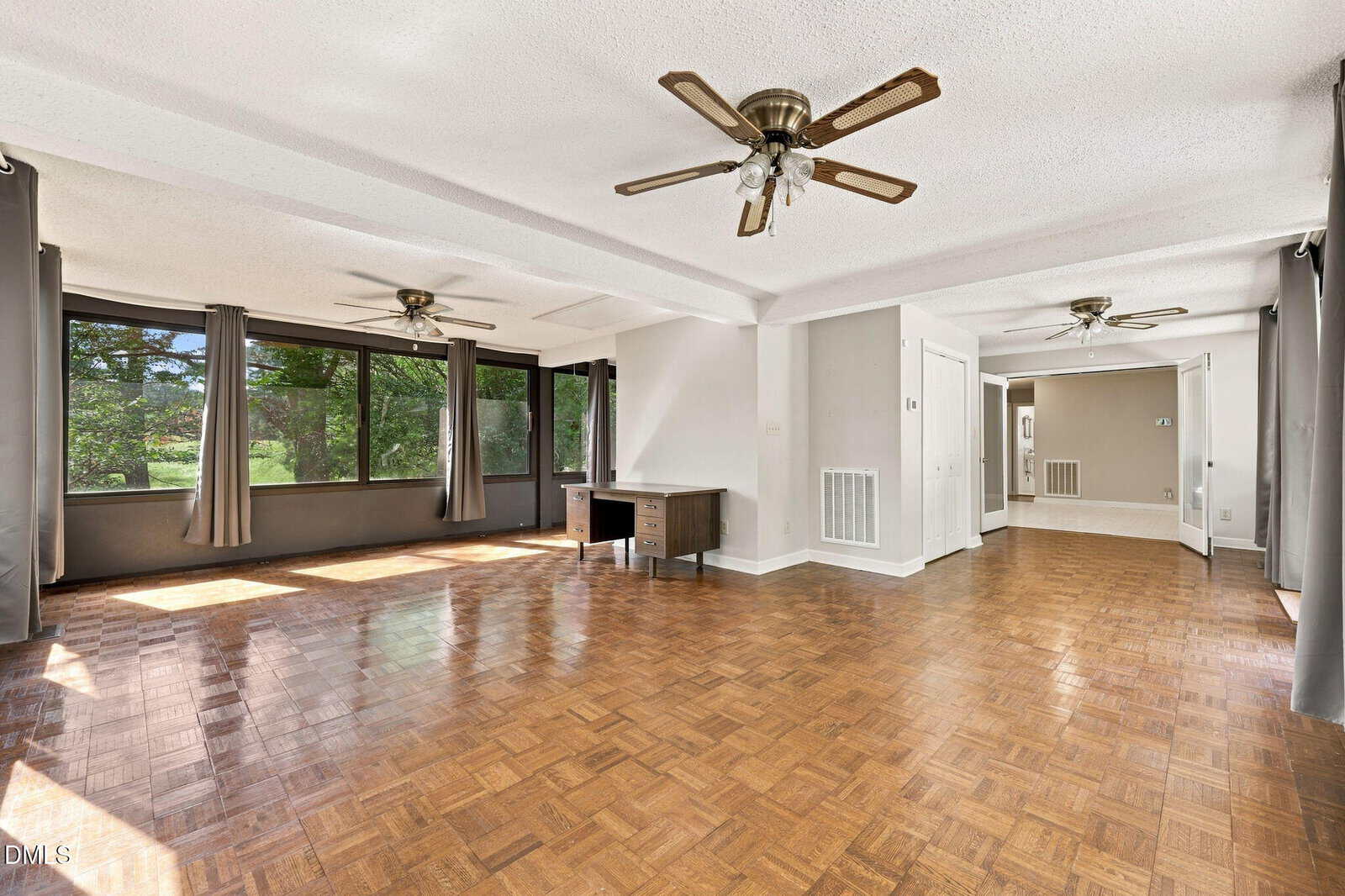 Spacious room with parquet flooring, large windows with gray curtains, three ceiling fans, white walls, and a small desk near the windows. Sunlight streams in, creating a bright and open atmosphere.