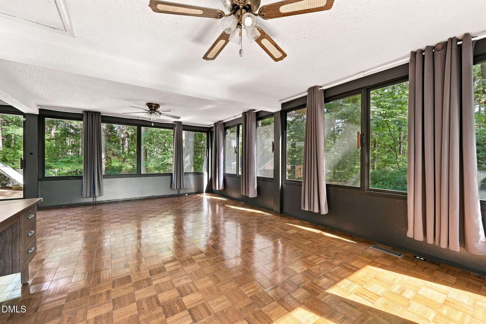 Spacious sunroom with parquet wood flooring, two ceiling fans, large windows with gray curtains on three sides, and a view of trees outside. The room is empty and has white ceilings with recessed lighting.