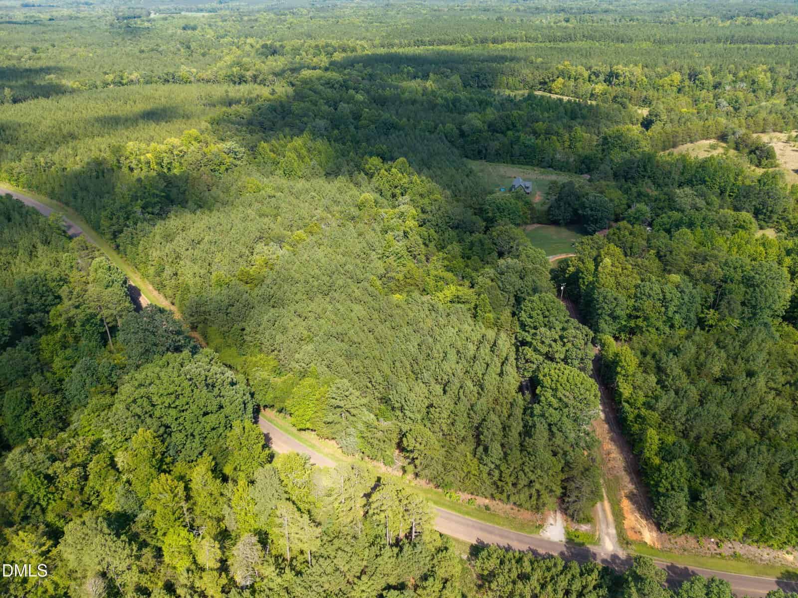Aerial view of a rural, wooded landscape featuring winding roads through dense green forest. A small clearing with a building is visible among the trees. The area is surrounded by lush vegetation.
