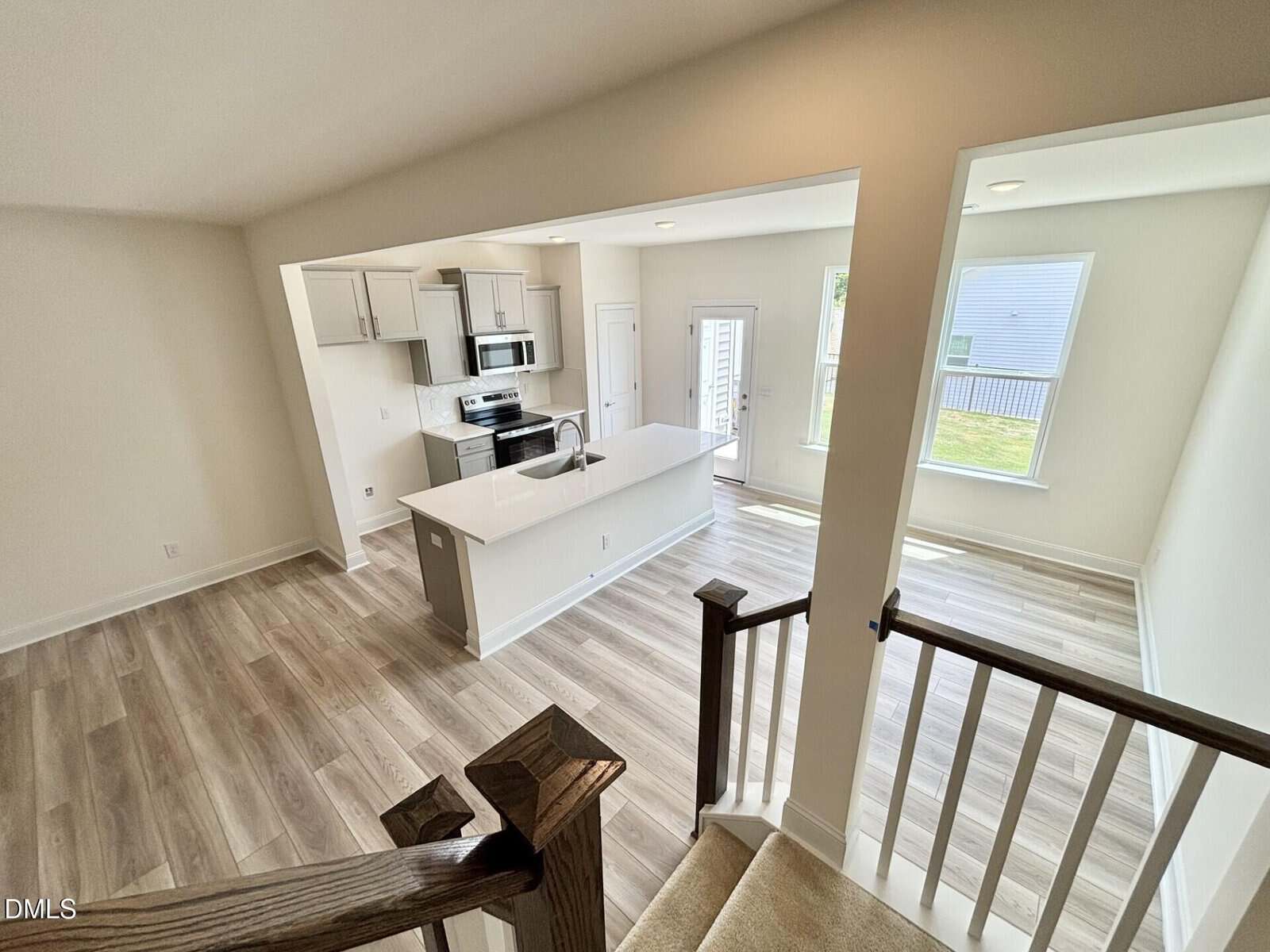 Modern kitchen with white cabinets, stainless steel appliances, and a central island, viewed from a staircase. The space features light wood flooring, large windows, and an open layout with plenty of natural light.