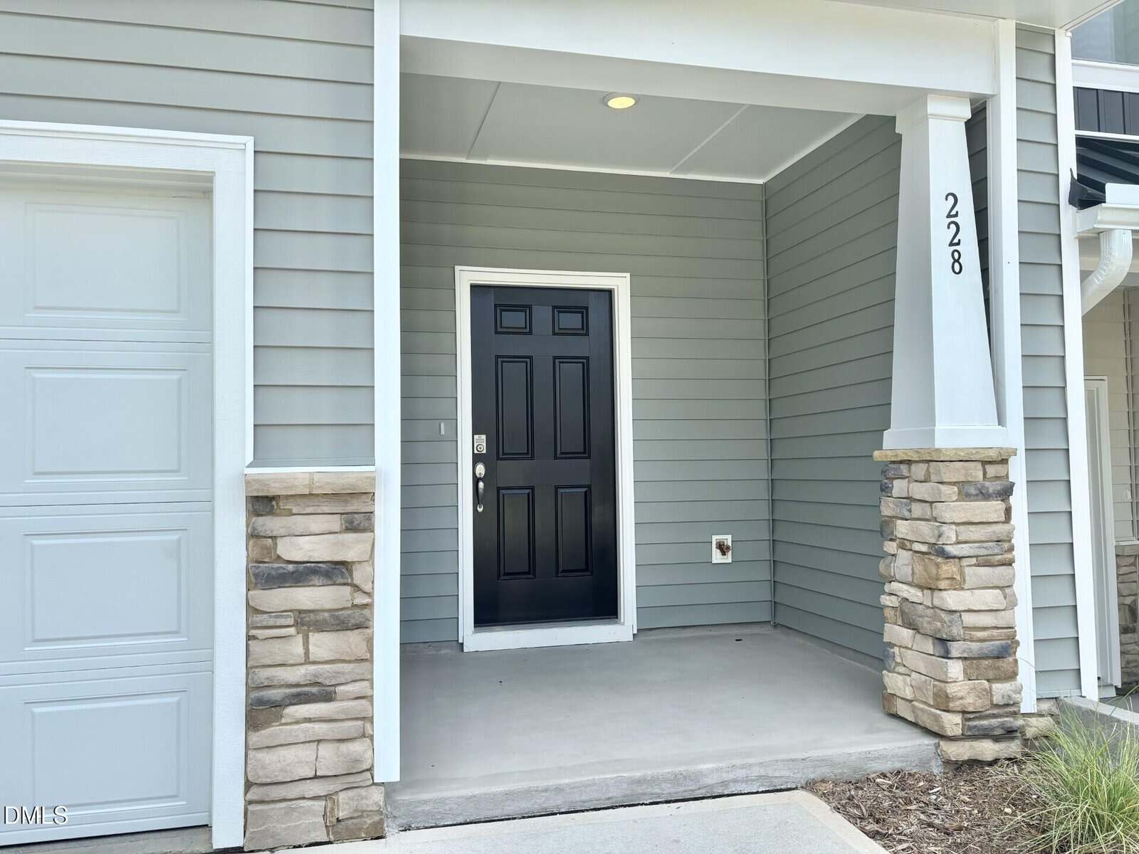 A front porch with gray siding and stone pillars, housing a black front door. To the left is a garage door, and the porch floor is concrete. The house number 228 is displayed on a white column.