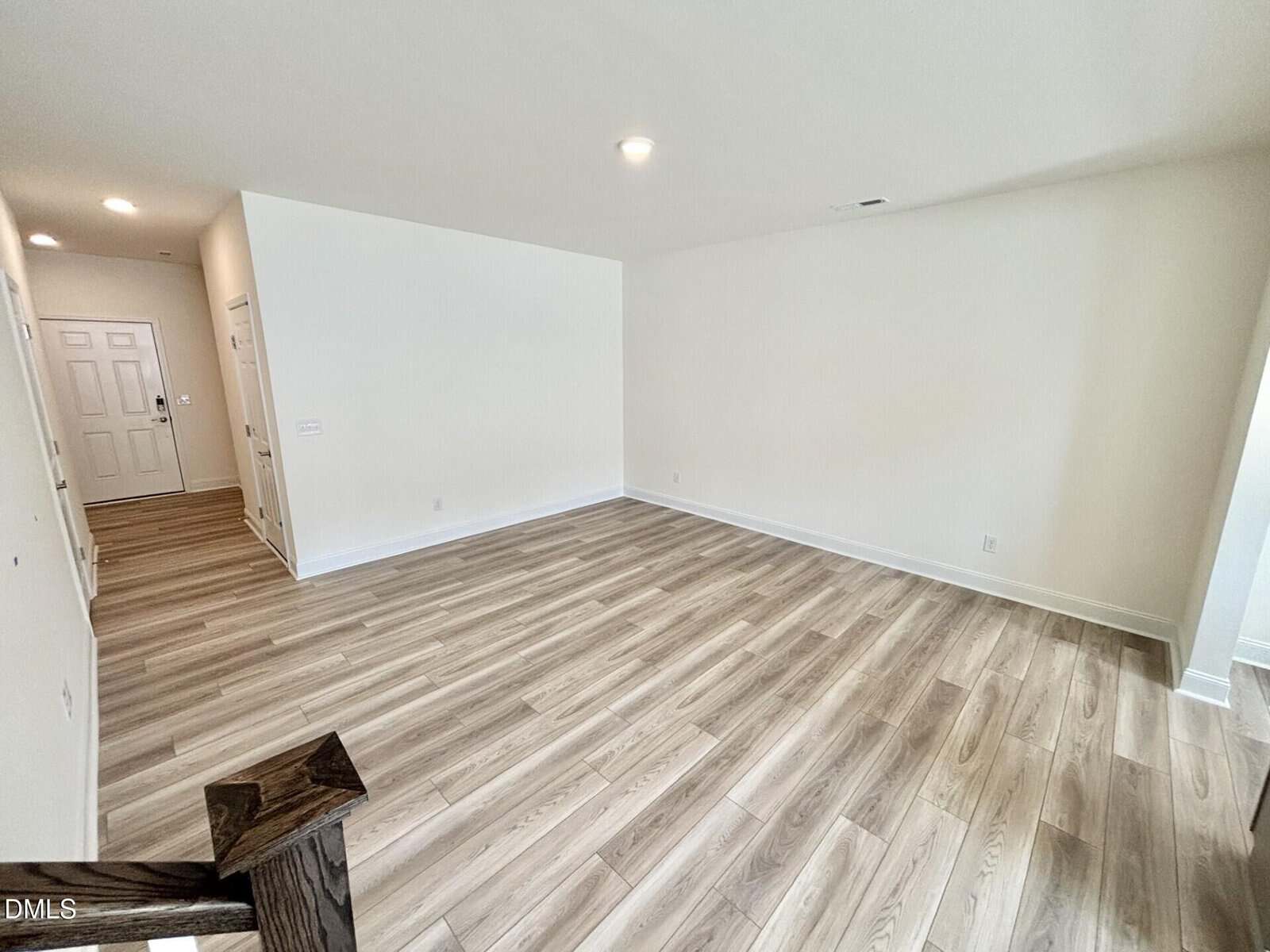 Spacious empty room with light wood flooring, white walls, and recessed ceiling lights. There’s a hallway leading to a white front door and visible stairs with a dark handrail in the foreground.