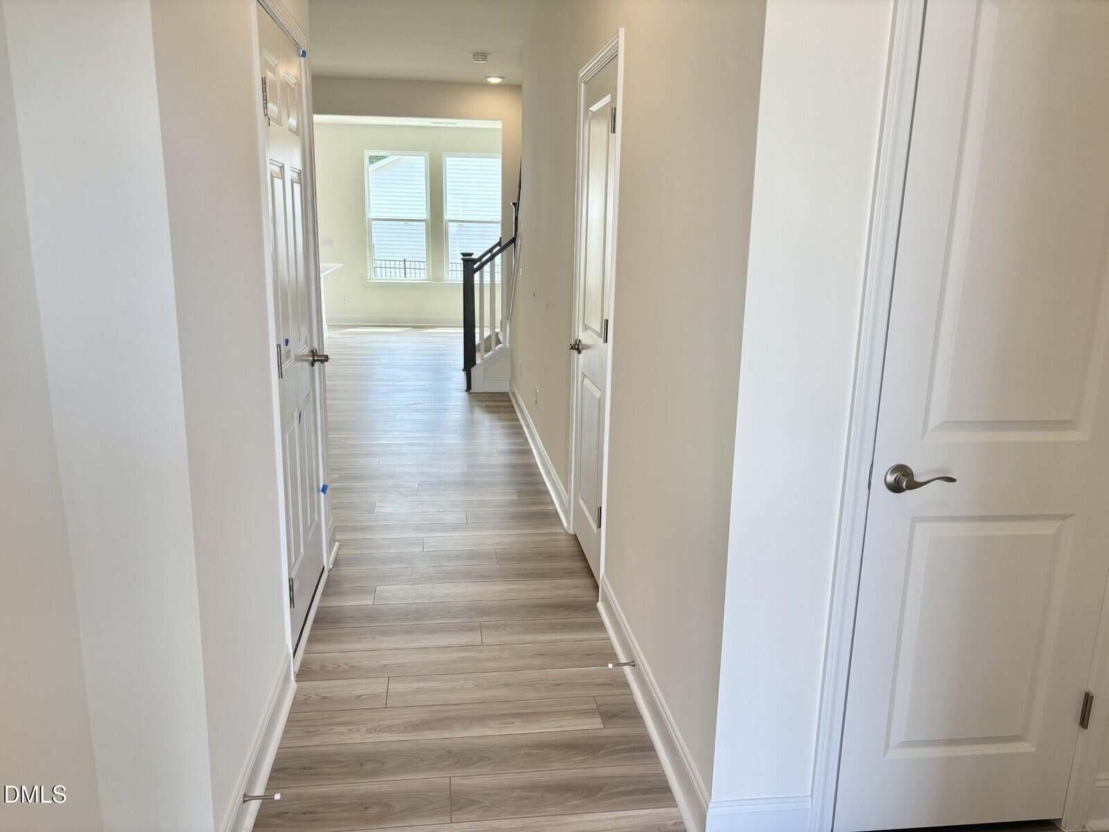 A hallway with light wood flooring, cream walls, four closed white doors, and a view of a window-lit room with a railing at the far end.