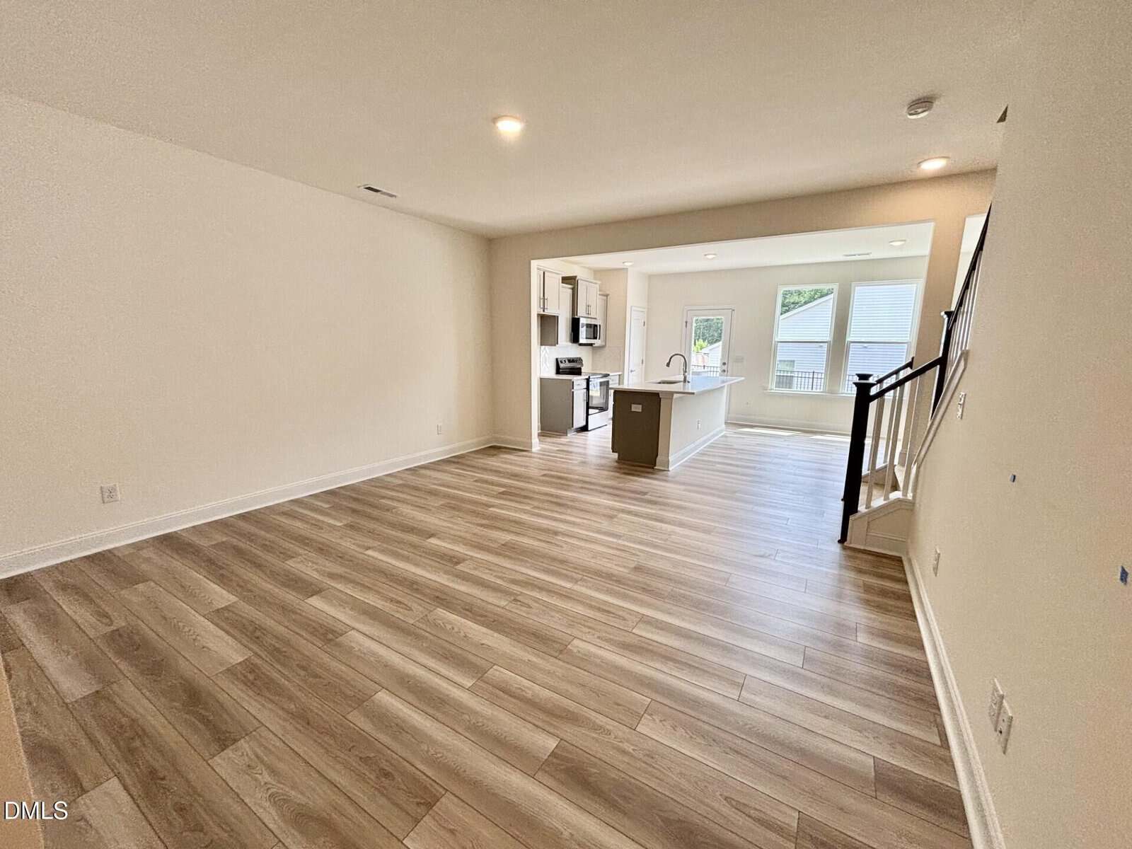 Open living area with light wood flooring, neutral walls, and recessed lighting. The space leads to a modern kitchen with white cabinetry, stainless steel appliances, an island, and a staircase on the right.