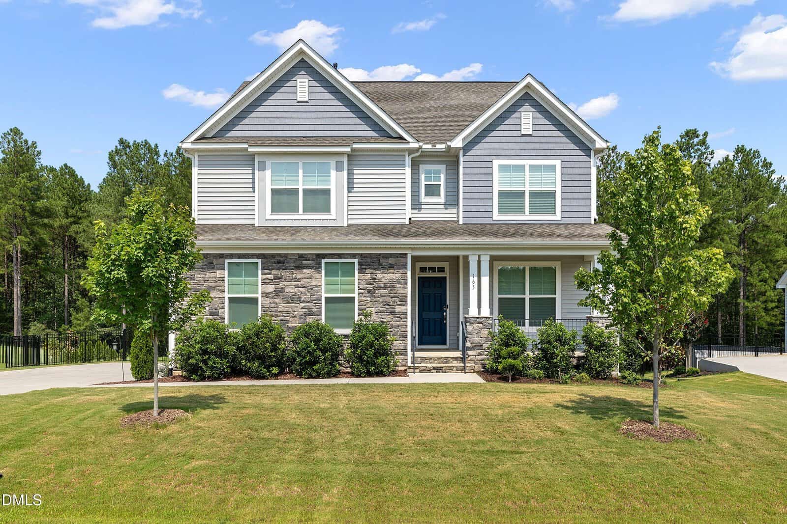 Two-story suburban house with gray siding, stone accents, and a blue front door. Well-kept front lawn, small trees, and bushes. Clear sky with some clouds and trees in the background.