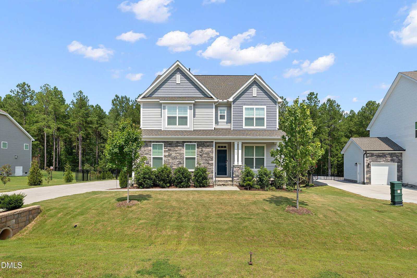 Two-story suburban house with gray siding and stone accents, a covered front porch, driveway, green lawn, small trees, and a wooded area in the background under a blue sky with scattered clouds.