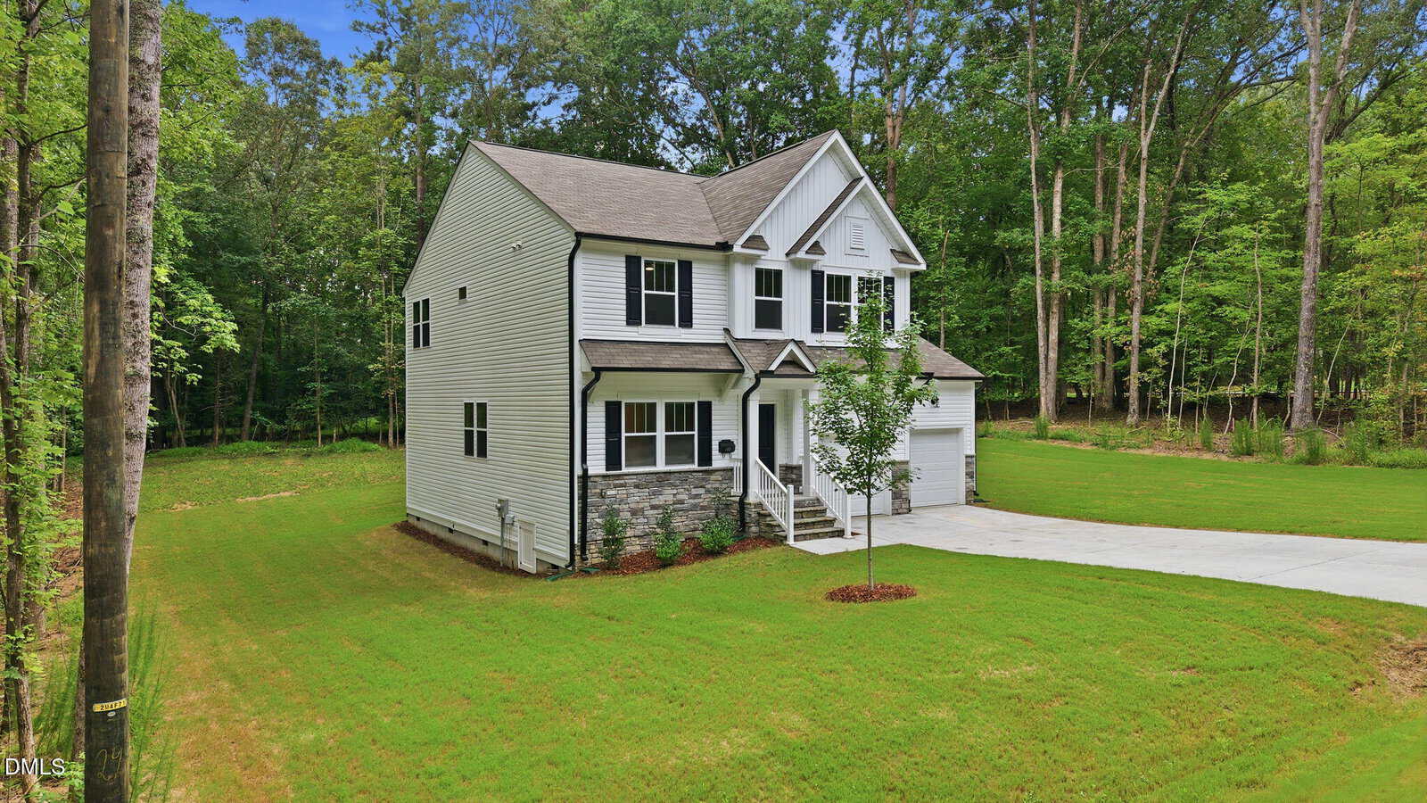 A two-story white house with black shutters and stone accents stands on a spacious green lawn, surrounded by tall trees. A driveway leads to a two-car garage attached to the right side of the house.