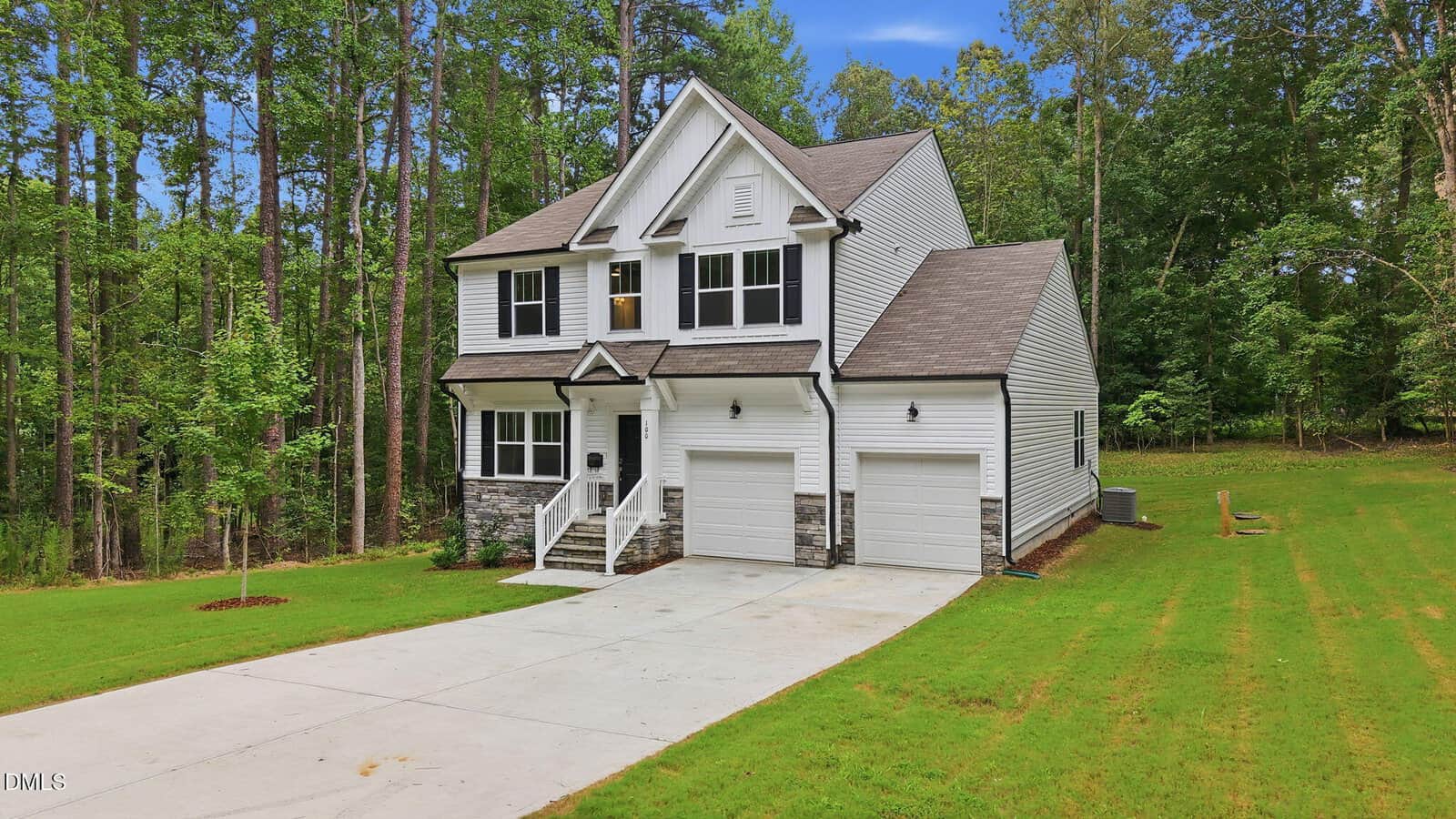 Two-story white house with gray shingles, a double garage, and stone accents on the lower facade. The home is surrounded by grass and trees, and there is a wide concrete driveway leading to the garage.