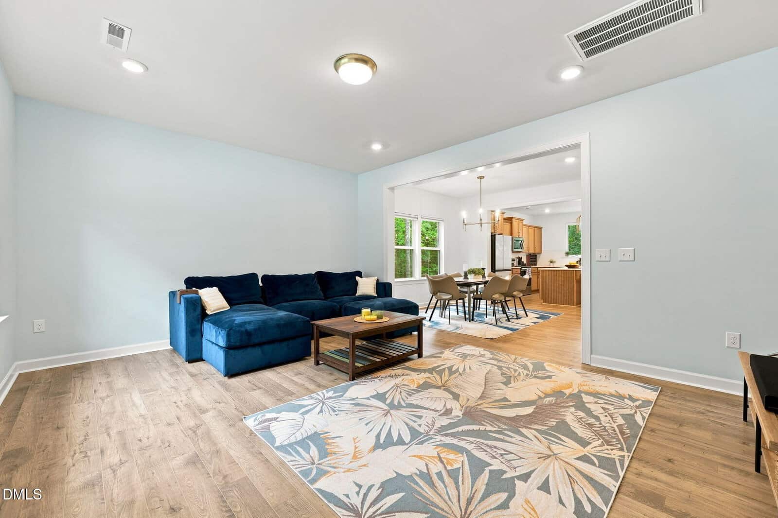 Open living room with a blue sectional sofa, wooden coffee table, and patterned rug, connected to a dining area and modern kitchen with light wood floors and pale blue walls.