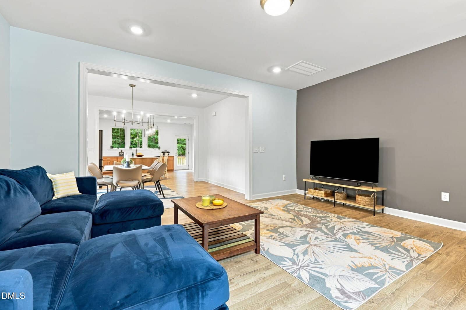 Modern living room with a blue sectional sofa, wooden coffee table, patterned area rug, and TV on a stand. An open doorway leads to a dining area and kitchen with wooden floors, white walls, and large windows.