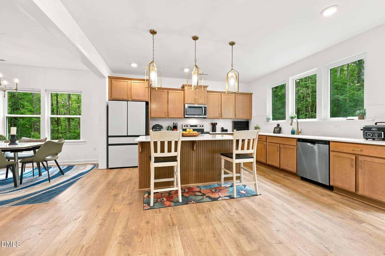 Modern kitchen with wood cabinets, island with two chairs, pendant lighting, stainless steel appliances, and large windows. Dining area with a table and chairs is visible on the left, with green trees seen through the windows.