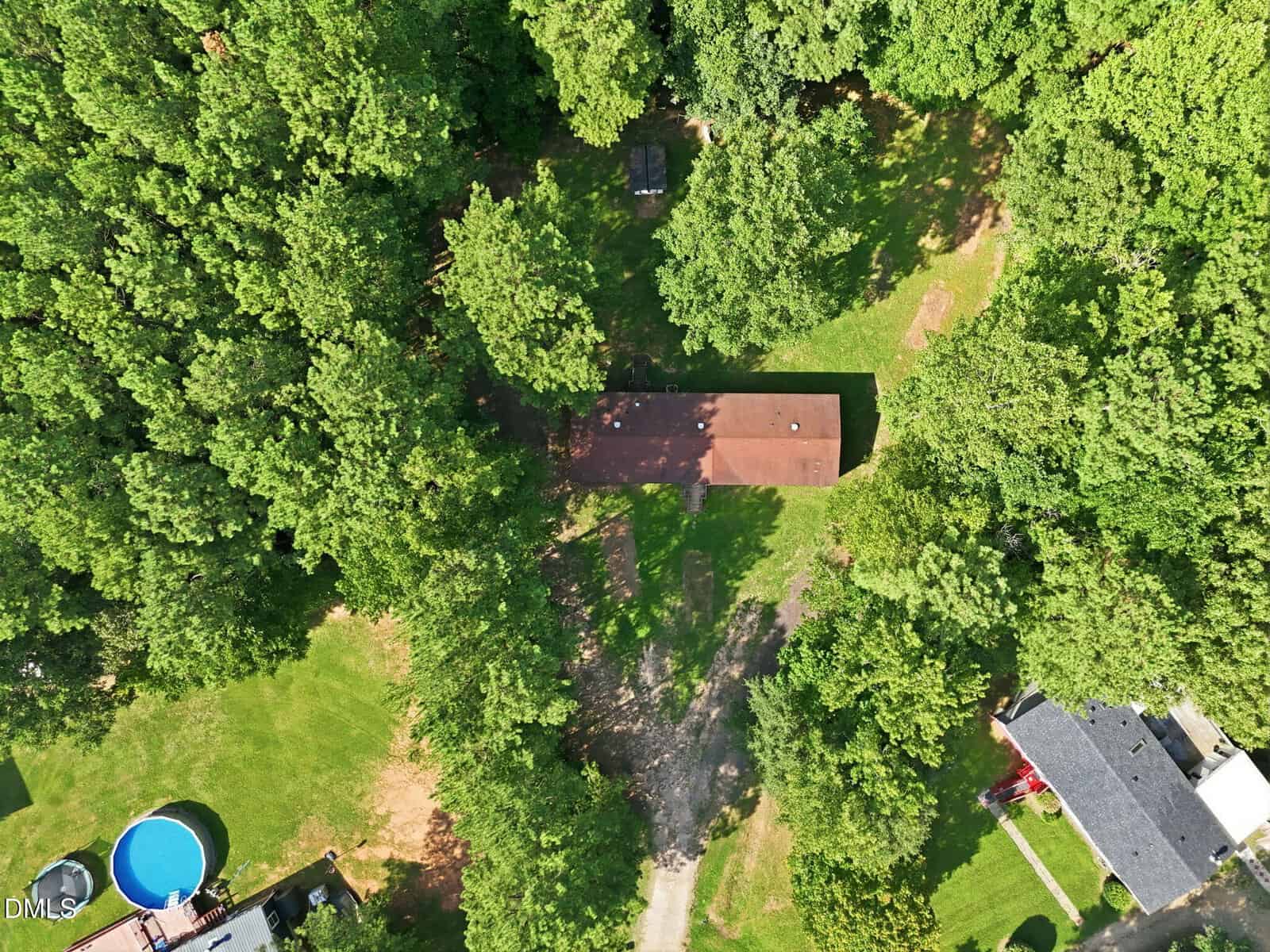 Aerial view of a rectangular brown-roofed house surrounded by dense green trees, with a driveway leading to it. A blue circular pool and adjacent houses are partly visible in the lower left and right corners.