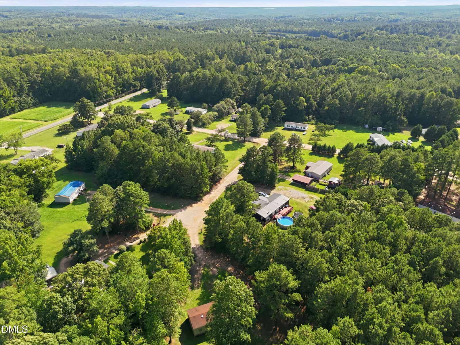 Aerial view of a rural neighborhood with houses, trees, a swimming pool, open grassy areas, and forested land stretching into the distance under a clear sky.