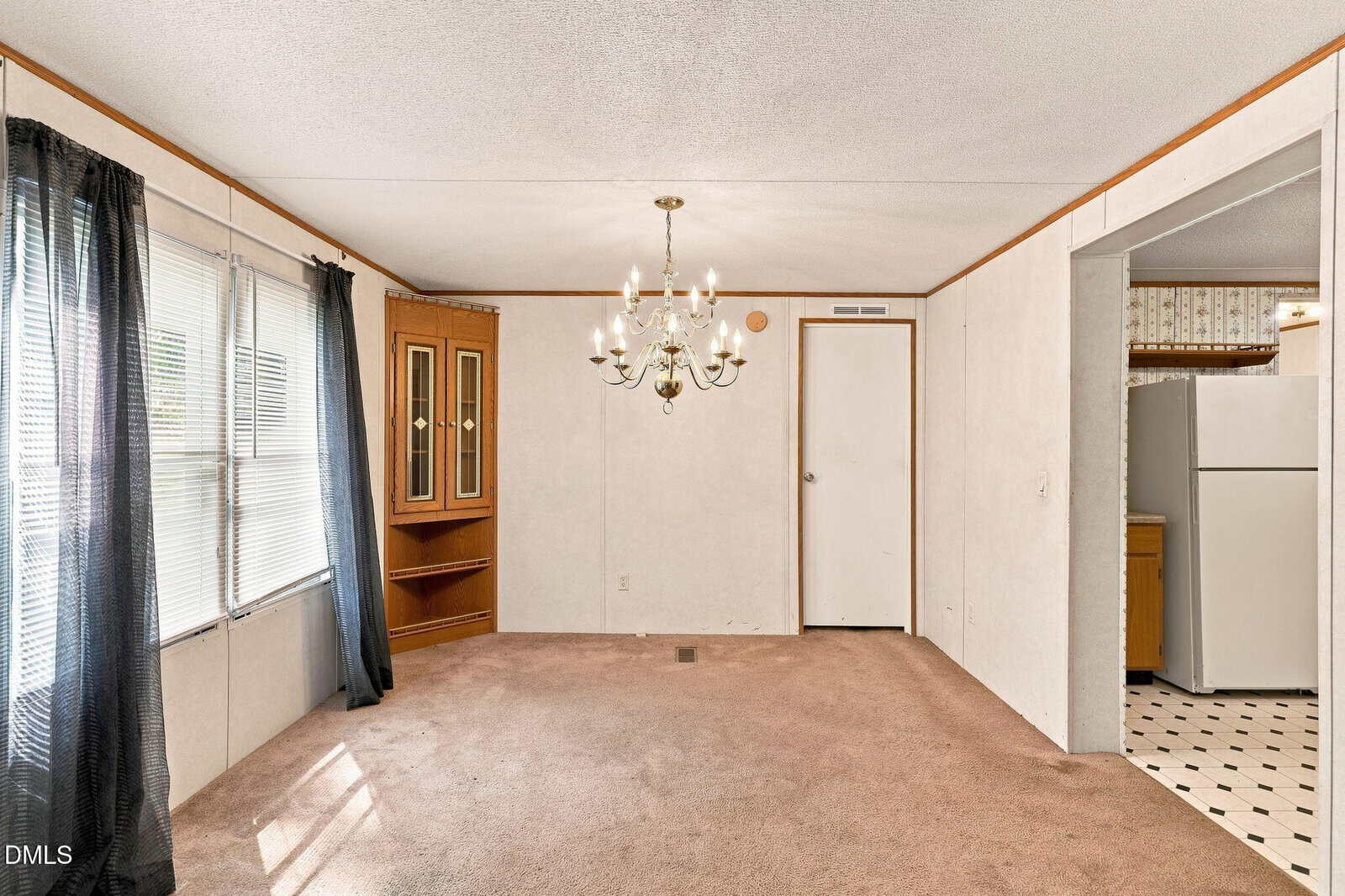 A carpeted living room with beige walls, a chandelier, corner china cabinet, and large windows with dark curtains; a doorway leads to a kitchen with white refrigerator and patterned linoleum floor.