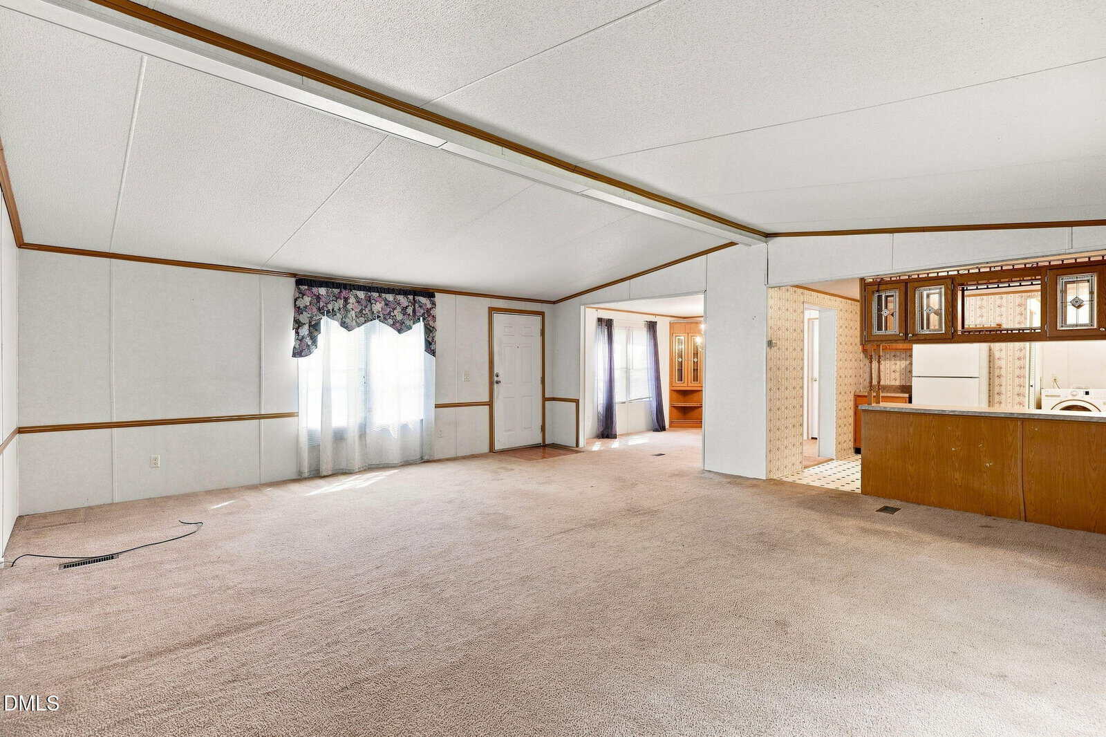 Spacious, carpeted living room in a manufactured home with white walls, wood trim, large window with floral valance, and view into kitchen area with brown cabinets and visible appliances.