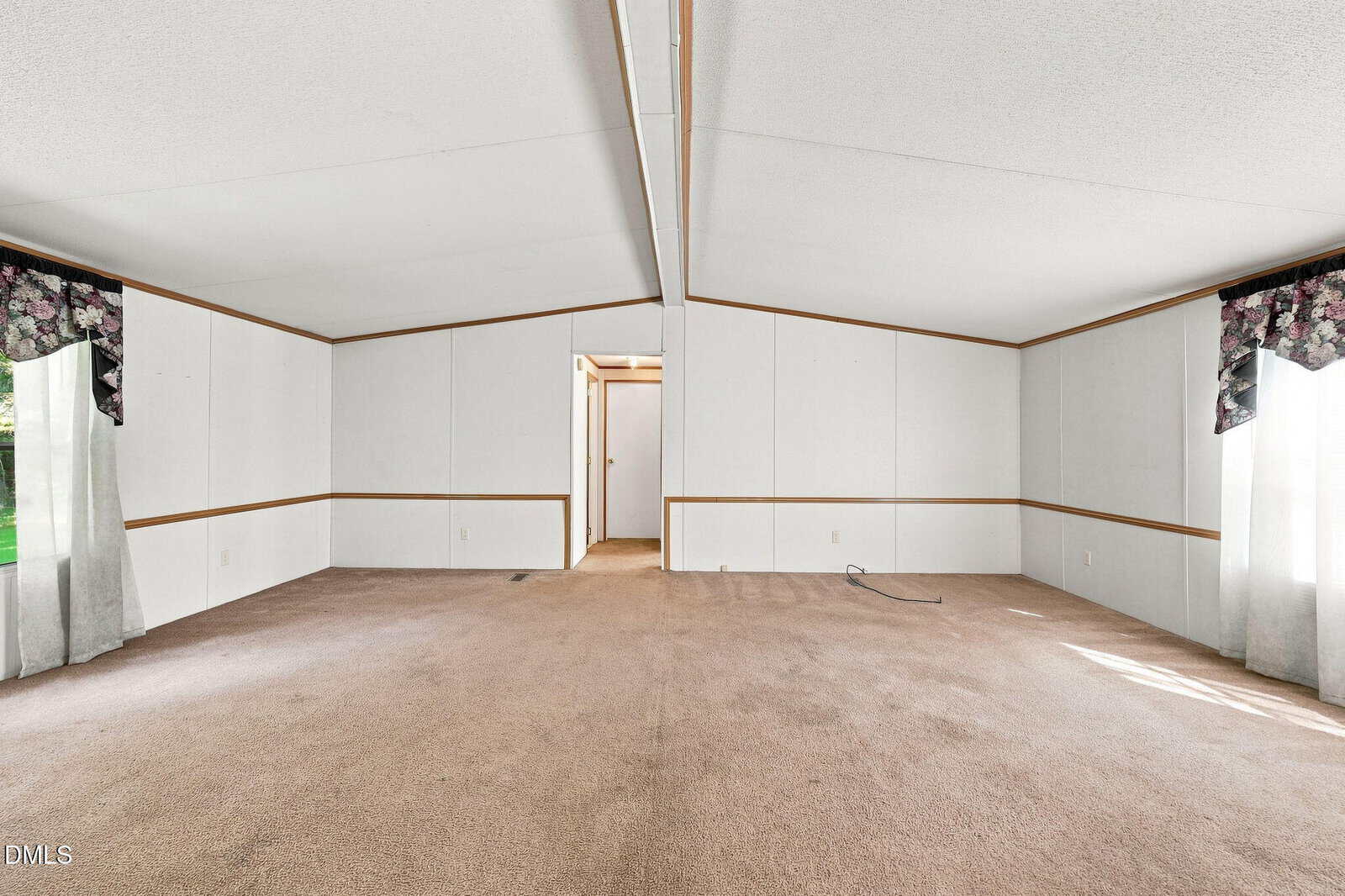 Empty room with beige carpet, white paneled walls with wooden trim, two windows with valances, and an open doorway leading to a hallway. Natural light enters from the windows.