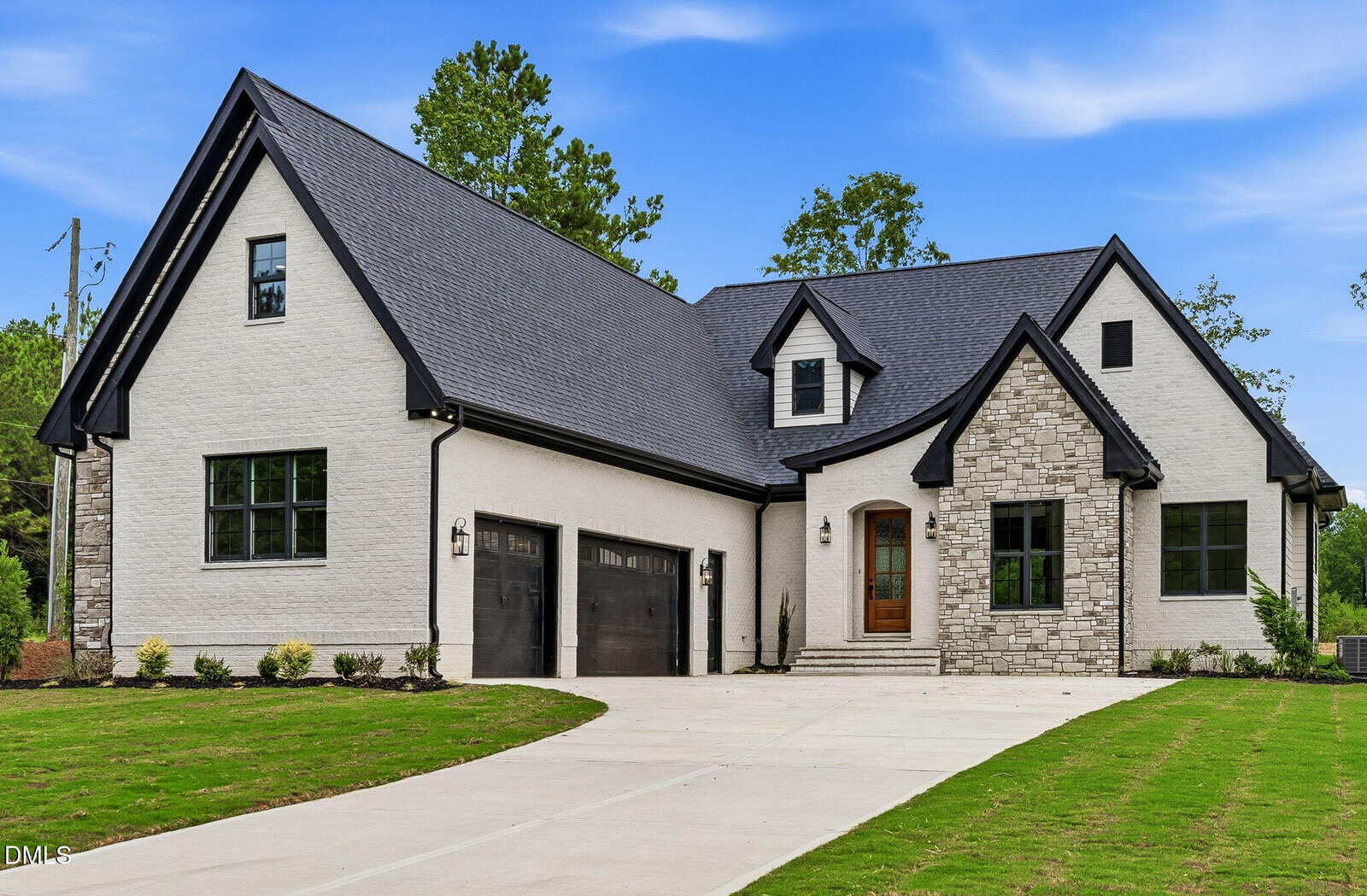 A modern two-story house with light brick and stone exterior, black roof, three black garage doors, a wooden front door, and a curved driveway, surrounded by green lawn and small shrubs.