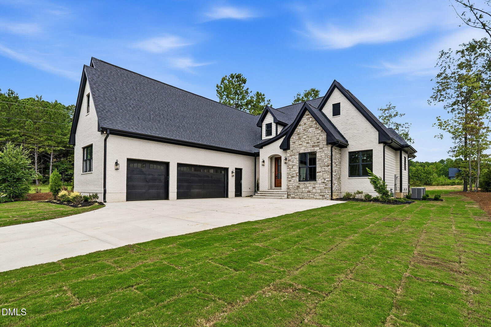 Modern two-story house with light brick exterior, dark roof, three-car garage, large driveway, and freshly planted front lawn. Trees and blue sky in the background.