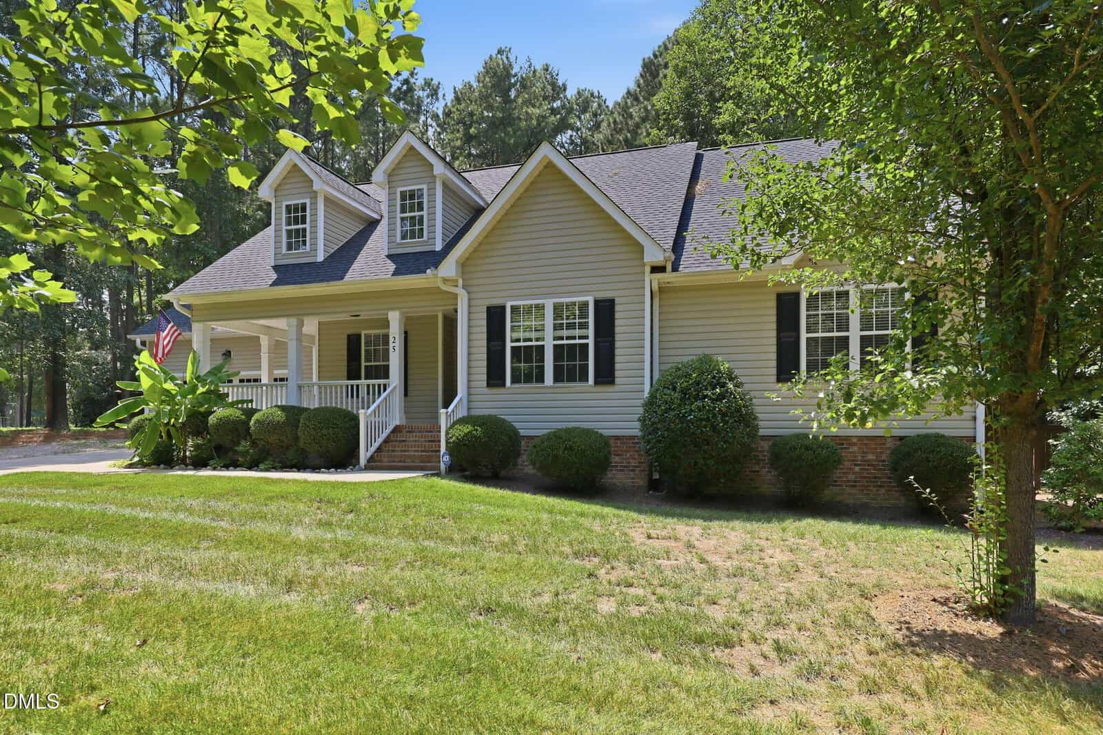 A single-story beige house with black shutters, a covered front porch, white railings, and dormer windows, surrounded by green grass, neatly trimmed bushes, and trees, with woods in the background.