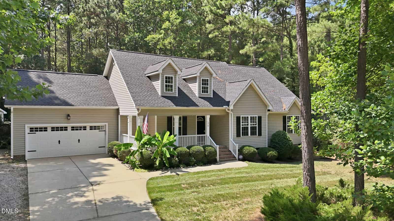 A light-gray house with white trim, an attached two-car garage, front porch with railings, dormer windows, and a concrete driveway, surrounded by green grass, bushes, and tall trees in the background.