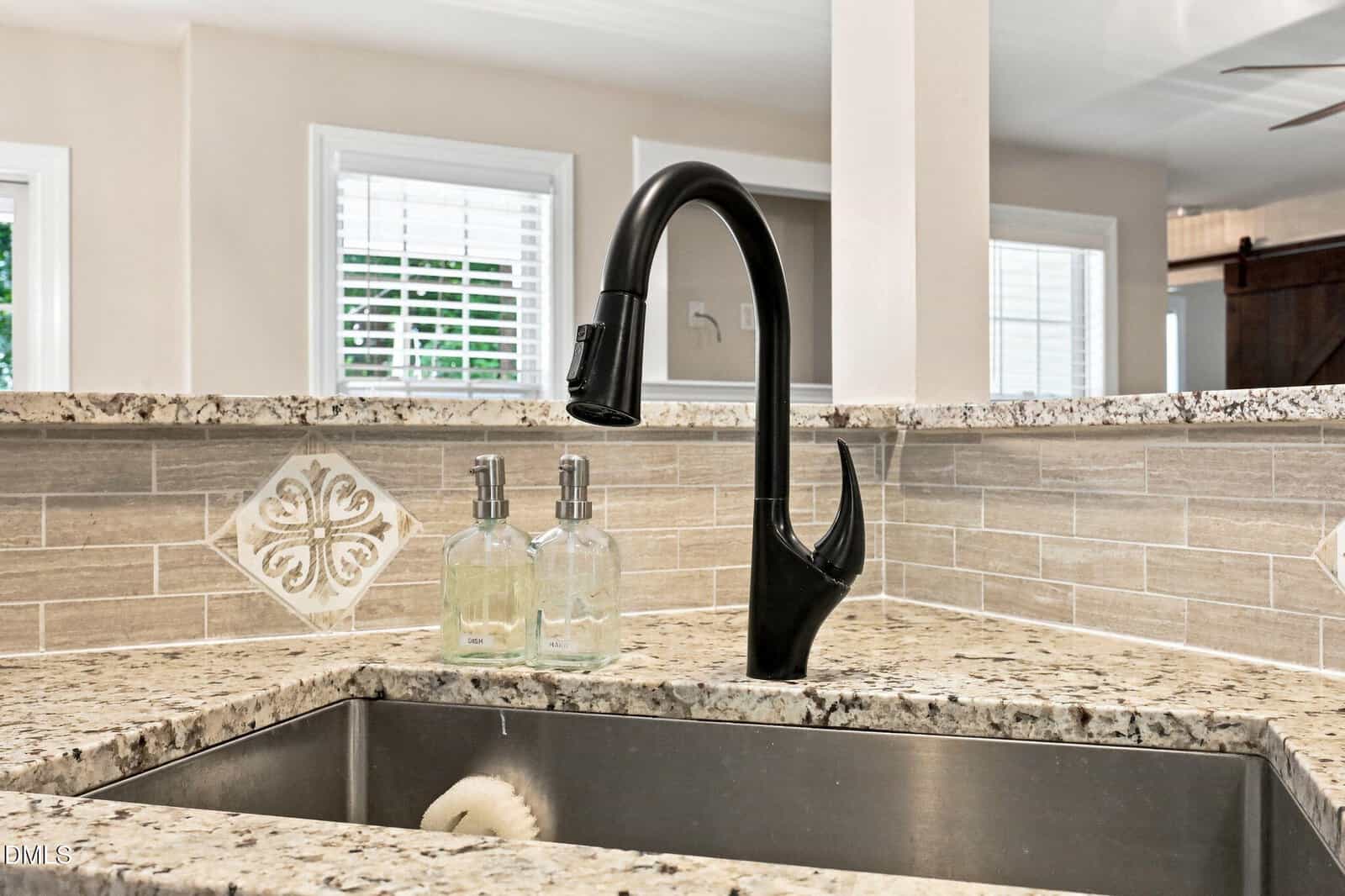 Modern kitchen sink with a black faucet and two soap dispensers on a granite countertop. Beige tile backsplash with a decorative tile accent is visible. Windows and part of the living space are in the background.