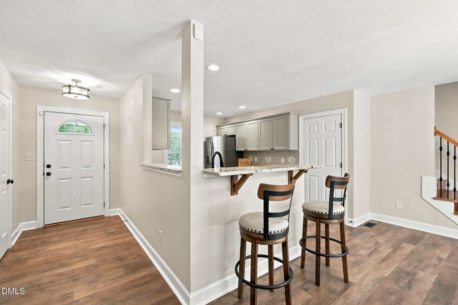 Modern kitchen with white cabinets, granite breakfast bar, and two cushioned barstools. Wood flooring, white walls, and an entry door with a window are visible. Stairs ascend to the right.
