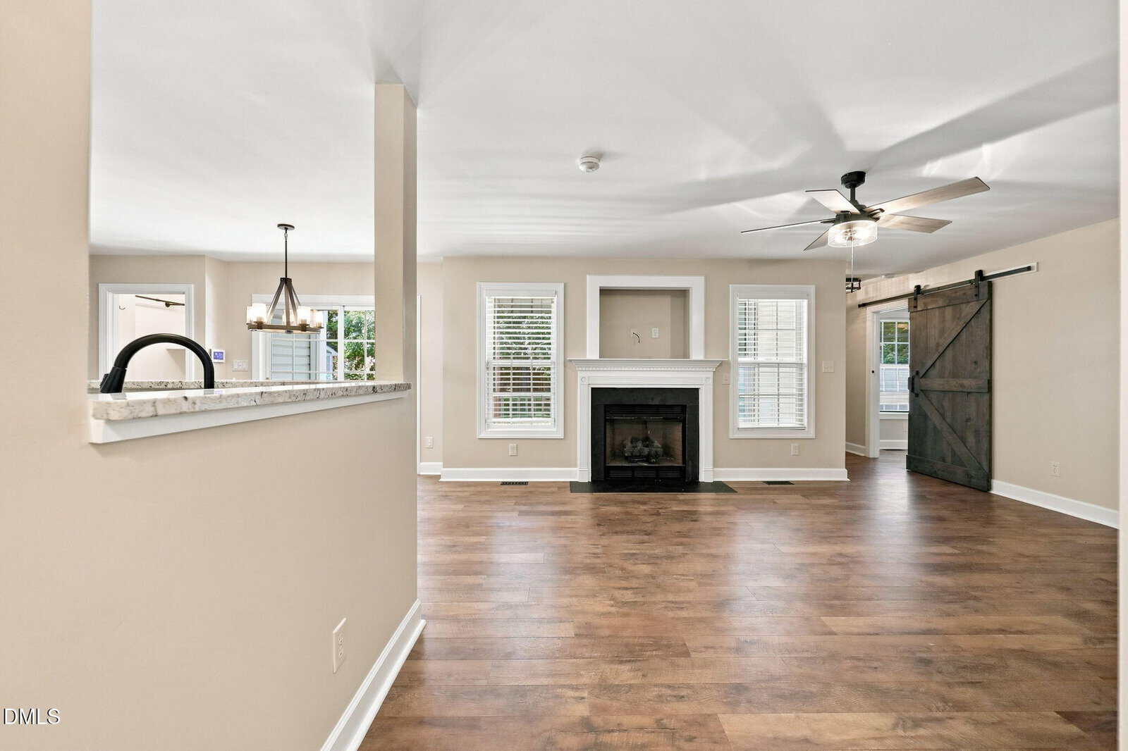 Open living room with wood floors, beige walls, a ceiling fan, fireplace, white shutters on windows, and a sliding barn door. Kitchen counter with a black faucet is visible on the left. Room is well lit with natural light.