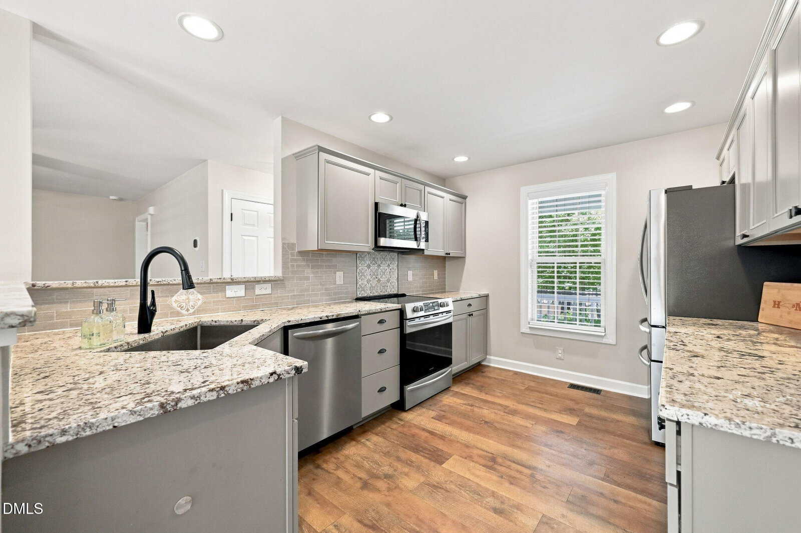 Modern kitchen with gray cabinets, stainless steel appliances, granite countertops, a tile backsplash, wood-style flooring, and a large window letting in natural light.