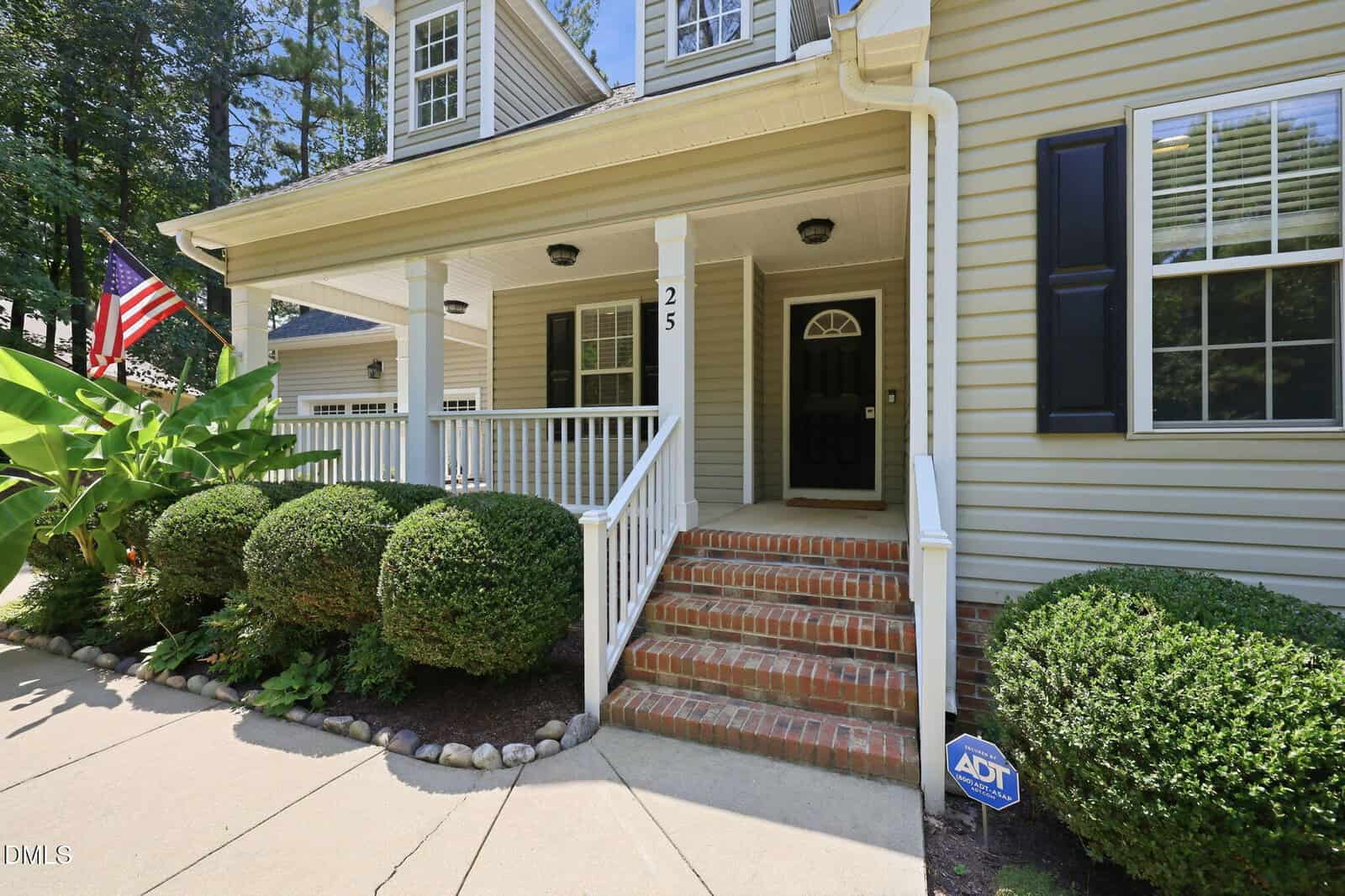 A house with yellow siding, black door, brick steps, white porch railing, well-kept bushes, an American flag, and an ADT security sign is shown on a sunny day. The house number 25 is visible by the front door.