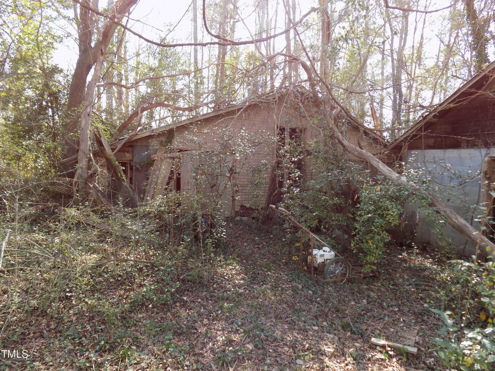 An abandoned, overgrown building surrounded by dense trees and vines, with sunlight filtering through the branches. Debris and a rusted machine are visible in the foreground.