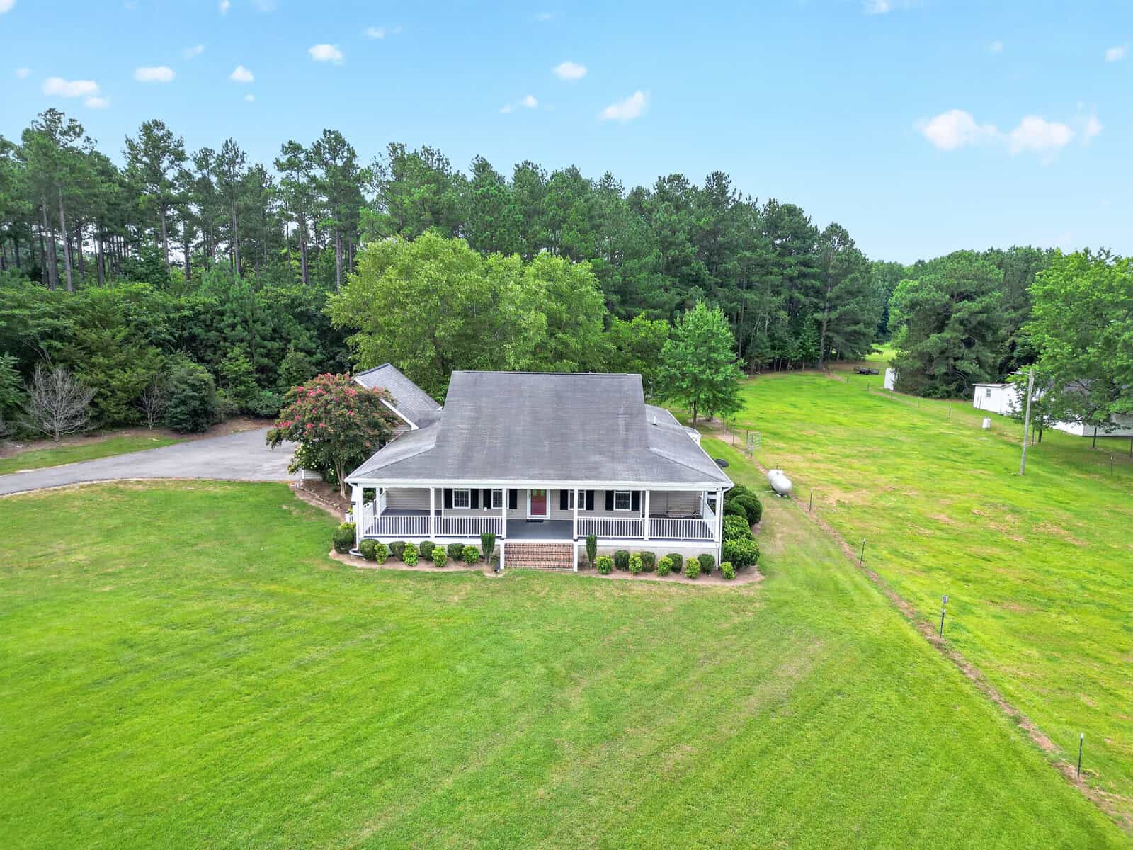 Aerial view of a country-style house with a wrap-around porch, centered in a large green grassy yard with a dense forest line in the background and a blue sky above.