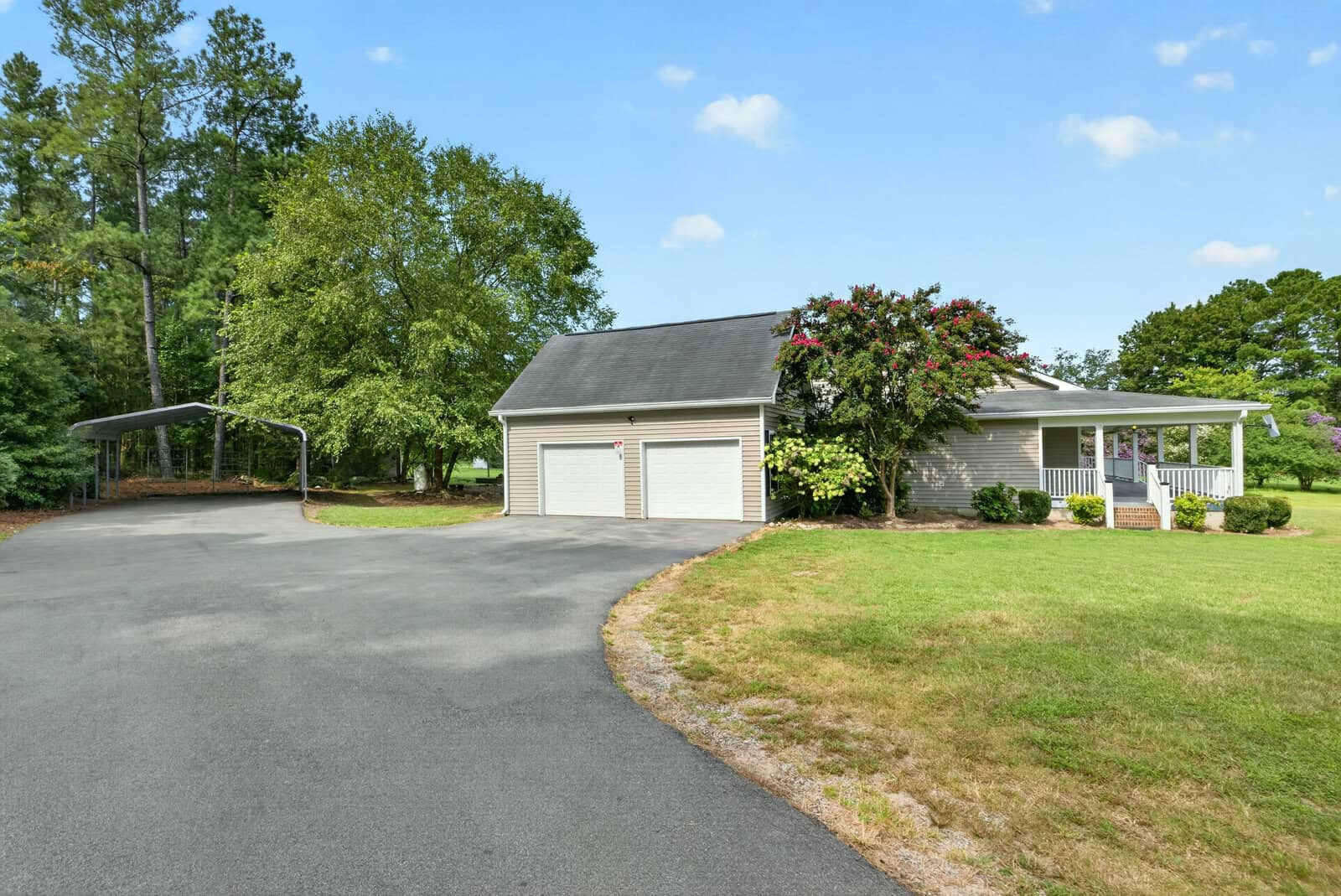 A small house with a front porch and two-car garage sits beside a paved driveway, surrounded by green grass, trees, and a covered carport on a clear day.