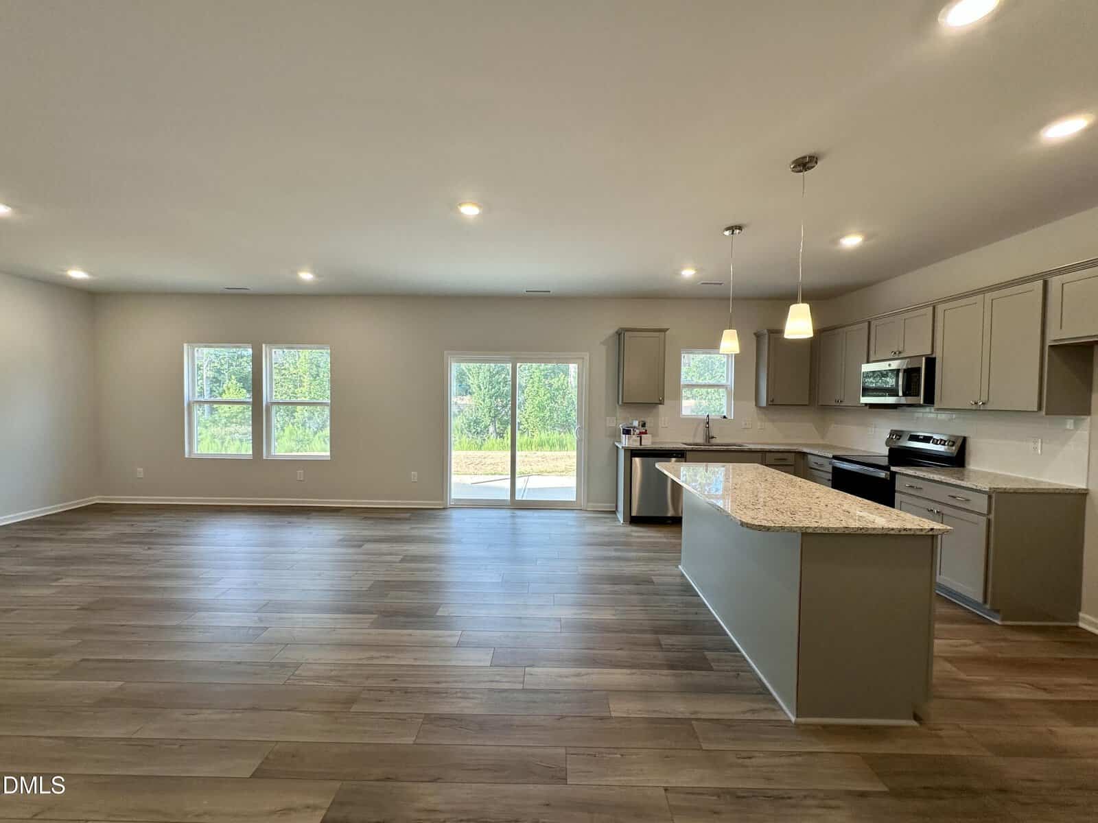 Modern open-concept kitchen and living area with wood floors, neutral walls, a large island, pendant lights, and stainless steel appliances. Sliding glass doors and windows let in natural light and overlook greenery.