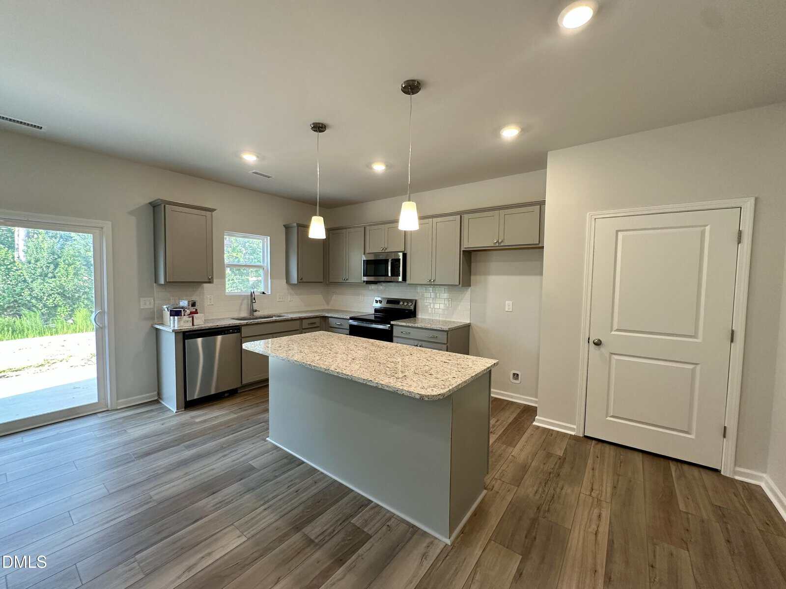 Modern kitchen with gray cabinets, stainless steel appliances, a central island with granite countertop, two pendant lights, wood-style flooring, and a door leading outside. The space is bright with natural and recessed lighting.