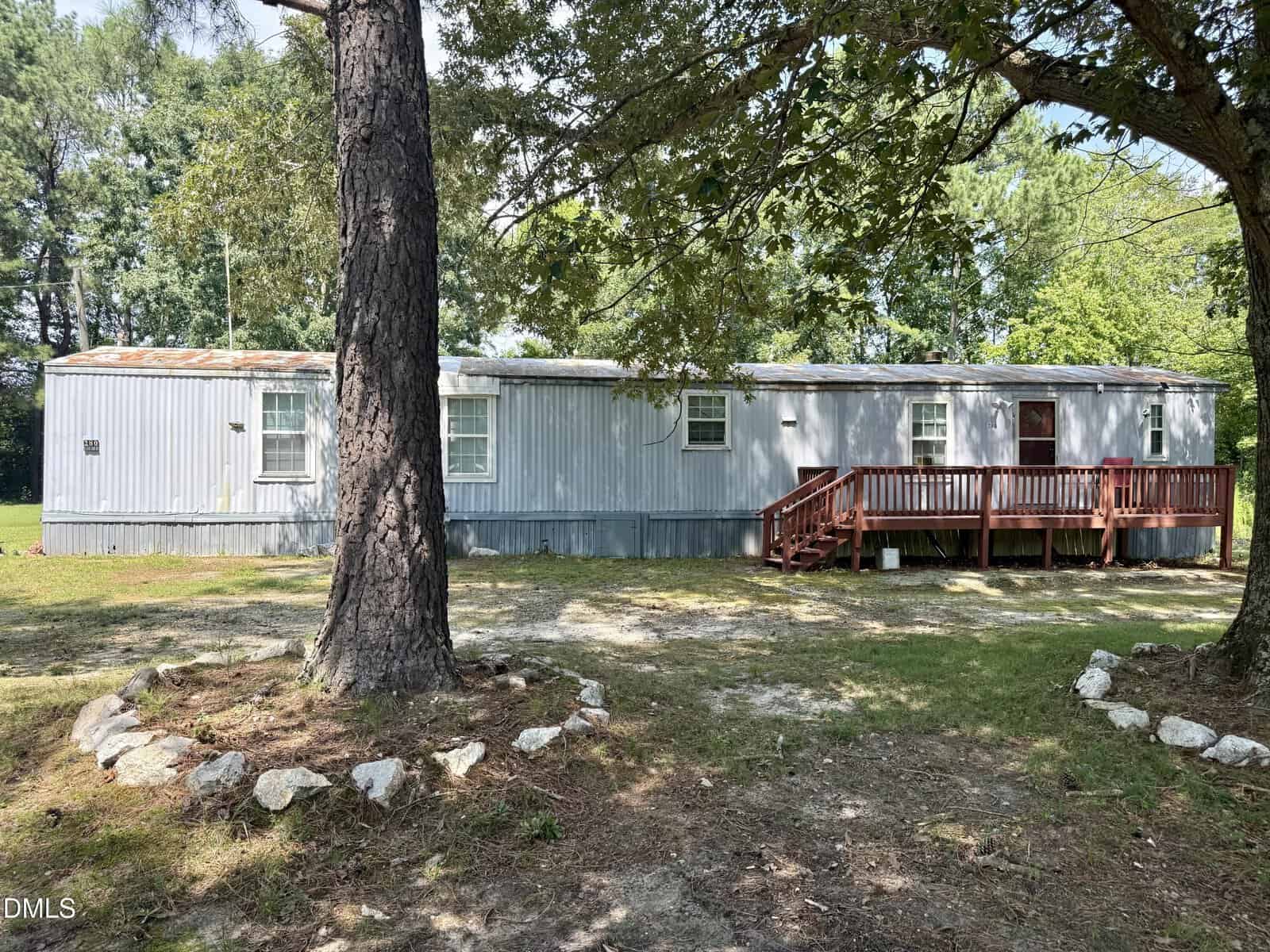 A long silver mobile home with a red wooden ramp sits among trees on a grassy yard at 280 Ellington Road, Manson. Sunlight filters through the branches, casting dappled shadows on the ground and the structure.