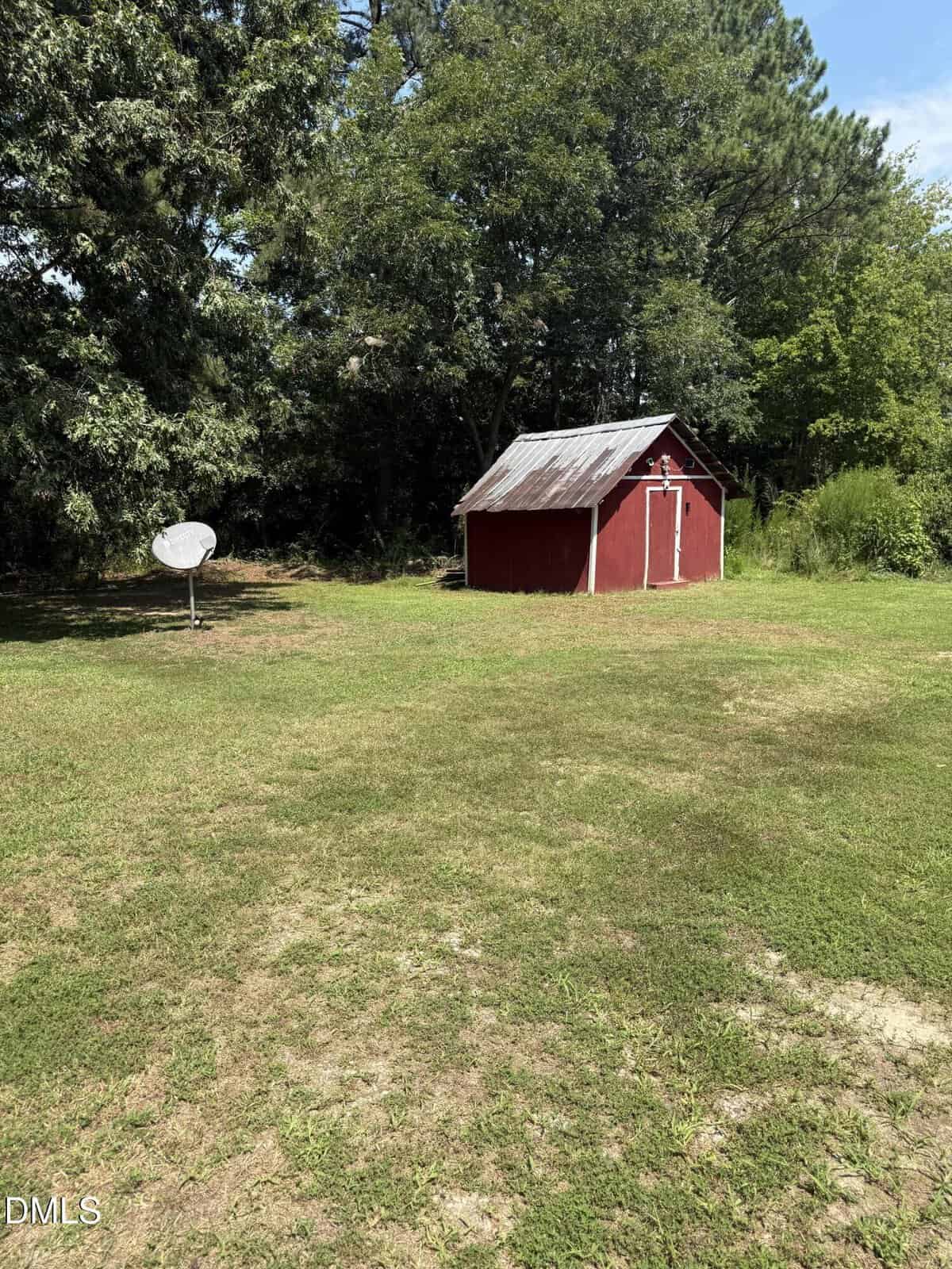 A small red shed with a metal roof sits in a grassy yard, surrounded by trees. A white satellite dish stands to the left of the shed. The sky is clear and sunny.