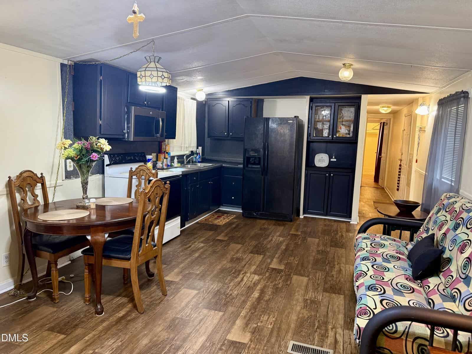 A cozy kitchen and dining area with dark cabinets, wood flooring, a small round table with two chairs, a floral arrangement, a patterned couch, and hallway leading to other rooms.