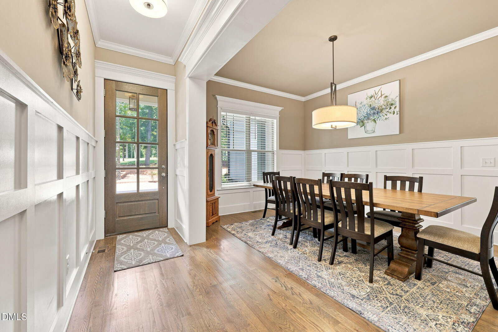 A dining room with a wooden table, ten chairs, a patterned rug, an overhead light, large windows with blinds, white wainscoting, and a front door with glass panels. A grandfather clock stands by the window.