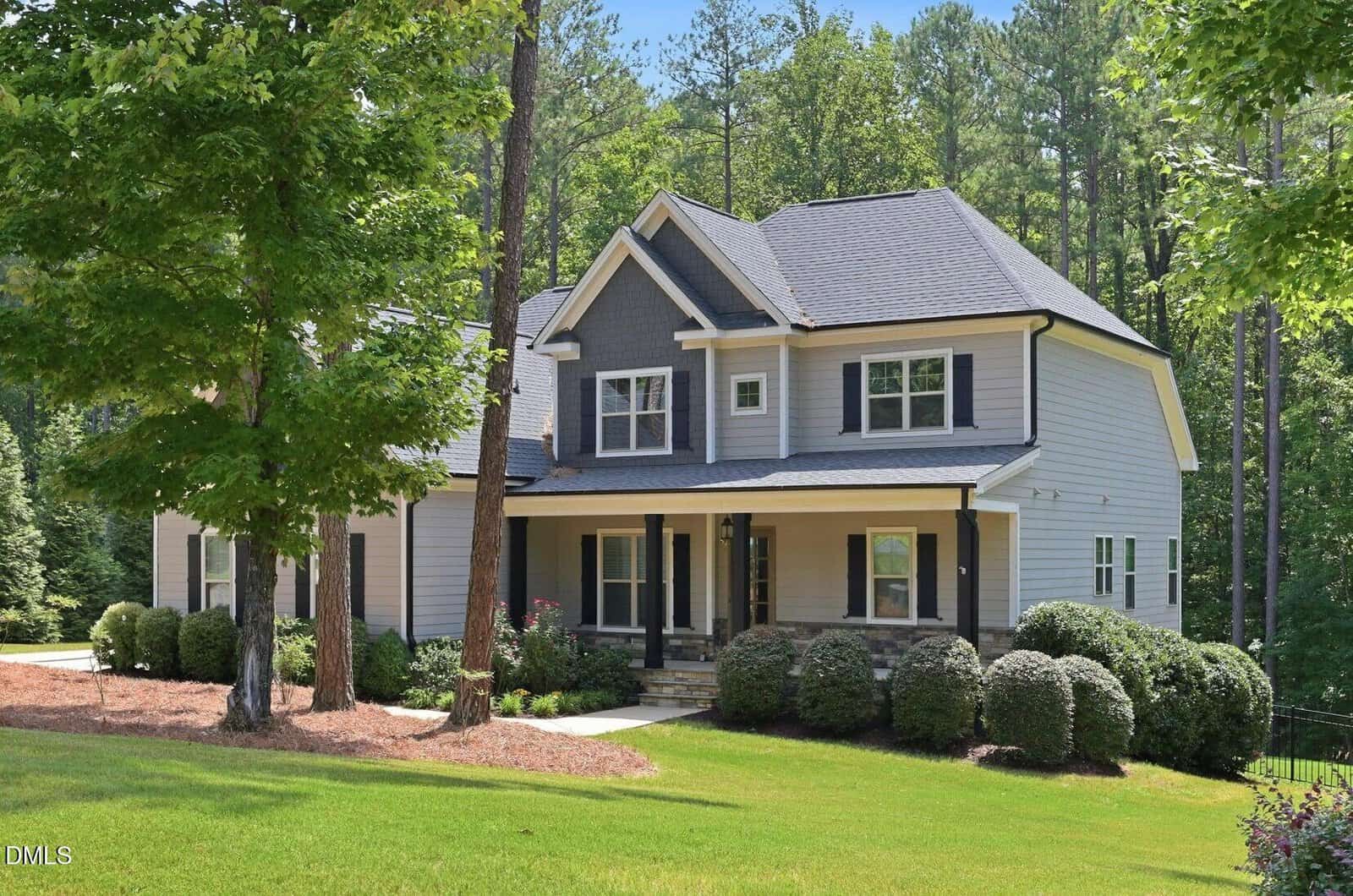 Two-story suburban house with gray siding, white trim, a covered front porch, and black shutters, surrounded by bushes, trees, and a well-maintained lawn on a sunny day.