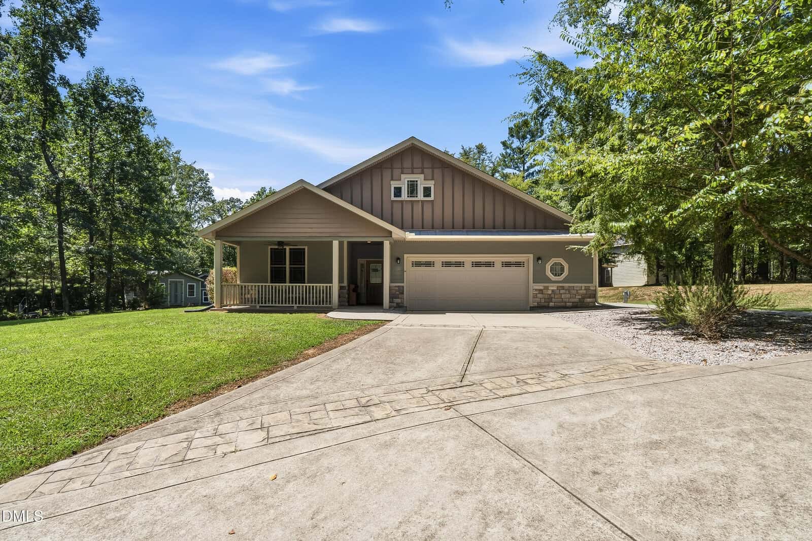 A modern single-story house with a front porch, double garage, and a wide driveway, surrounded by green lawn and trees under a clear blue sky.