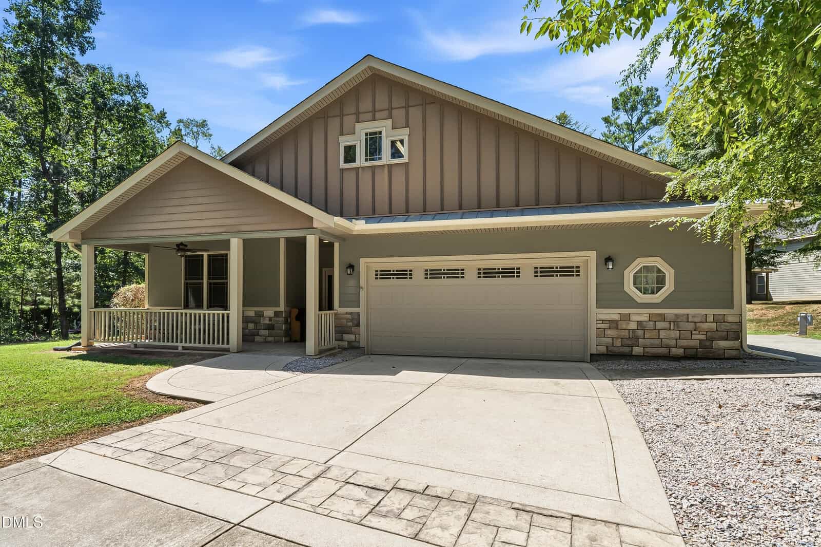 Single-story house with a covered front porch, double garage, grey siding, stone accents, and a driveway. Surrounded by trees, under a blue sky with light clouds.