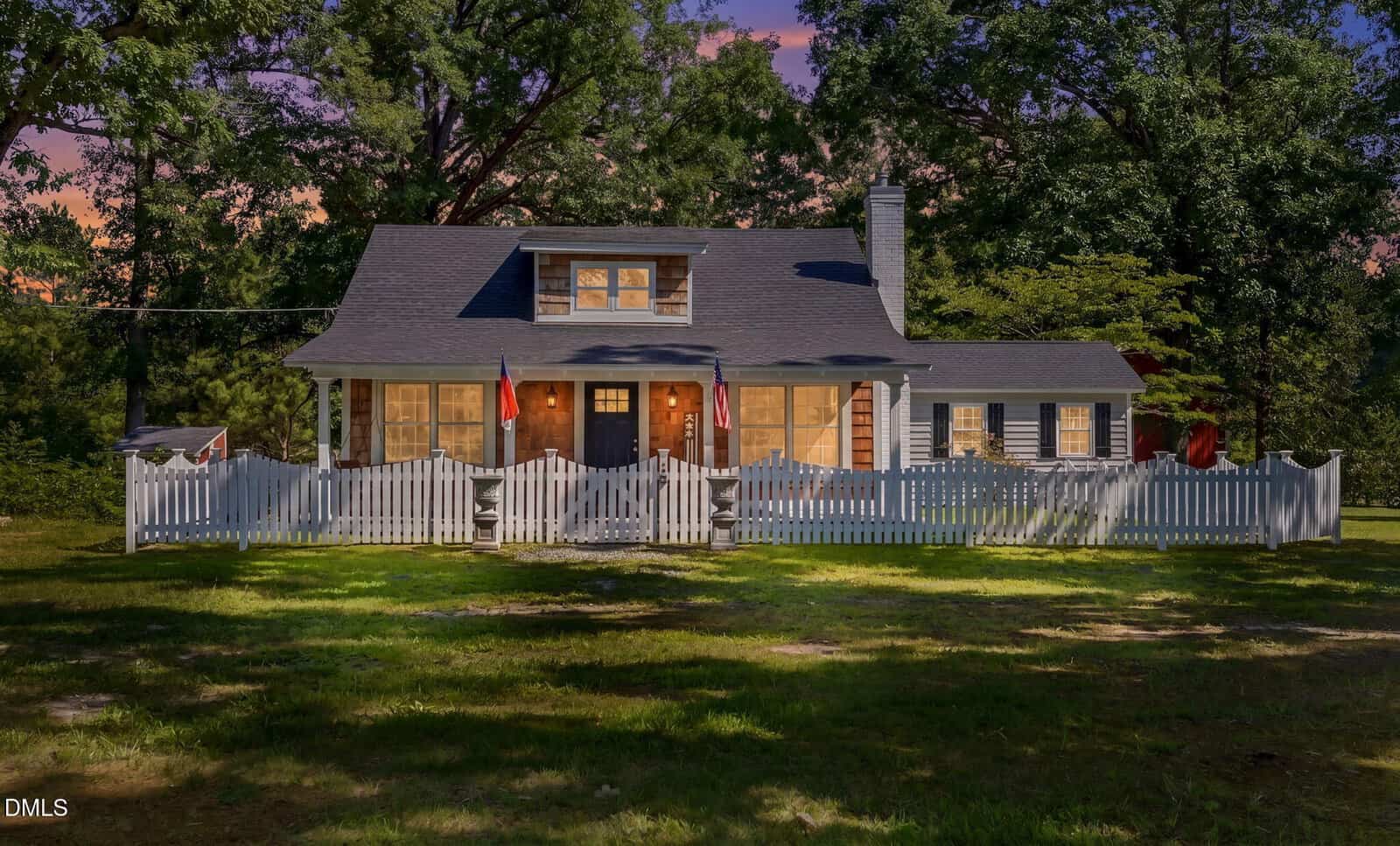 Charming white house with a front porch, white picket fence, and two red flags. Surrounded by trees, the house is lit warmly at dusk with a well-kept lawn in front.