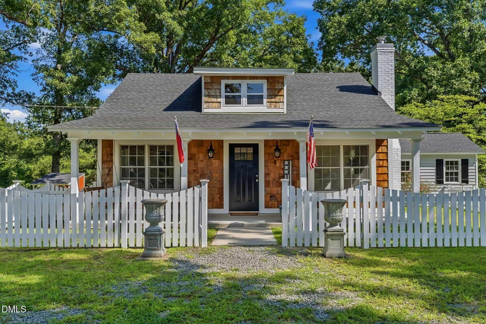 A charming cottage with brown shingles, white trim, and a black front door. Two flags hang at the front, with a white picket fence, urn planters, and a green yard surrounded by trees under a blue sky.