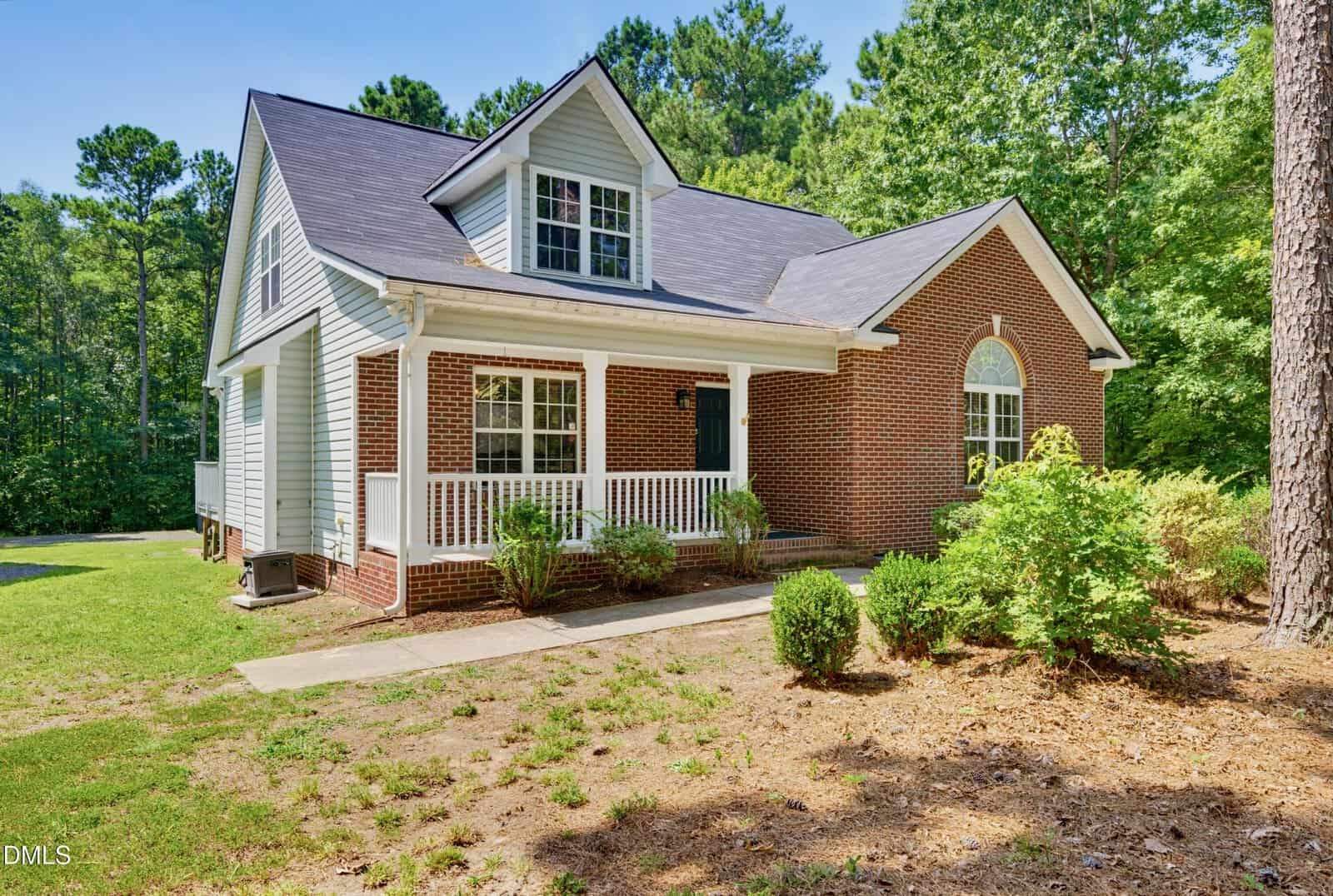 A one-and-a-half story house with brick and light blue siding, white trim, a covered front porch, and a gabled roof, surrounded by trees and greenery on a sunny day—this inviting residence is nestled at 1110 Lake Ridge Drive in the charming community of Creedmoor.