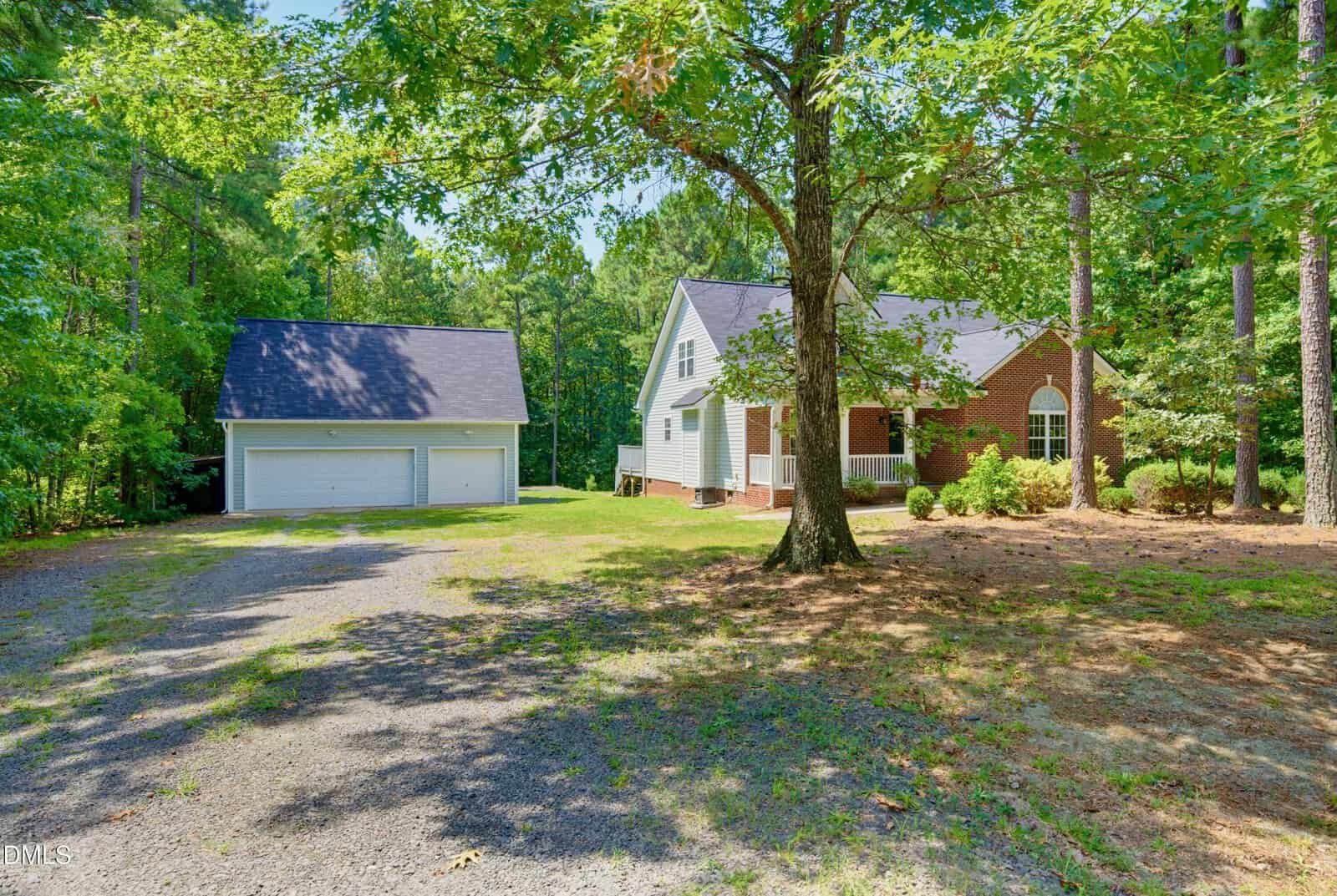 A brick house with white trim sits among tall trees, with a detached two-car garage and a gravel driveway leading up to both buildings on a sunny day.