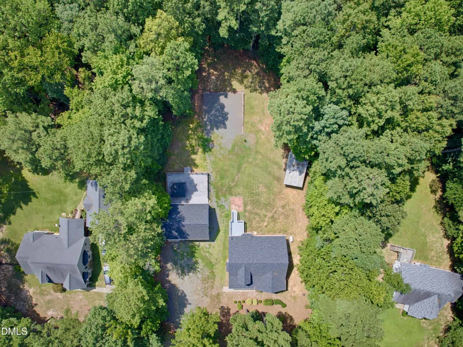 Aerial view of several houses surrounded by dense green trees, with driveways and small yards visible between the homes. One property includes a small shed and open grassy area.
